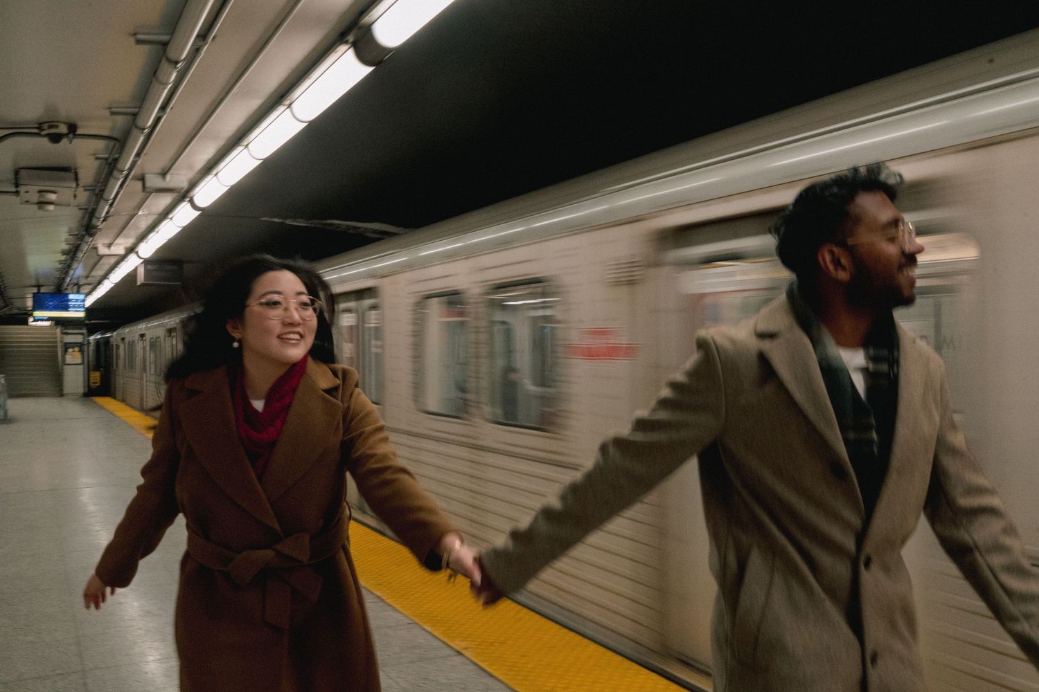 Two people hold hands and run playfully along a subway platform as a train passes by in the background.