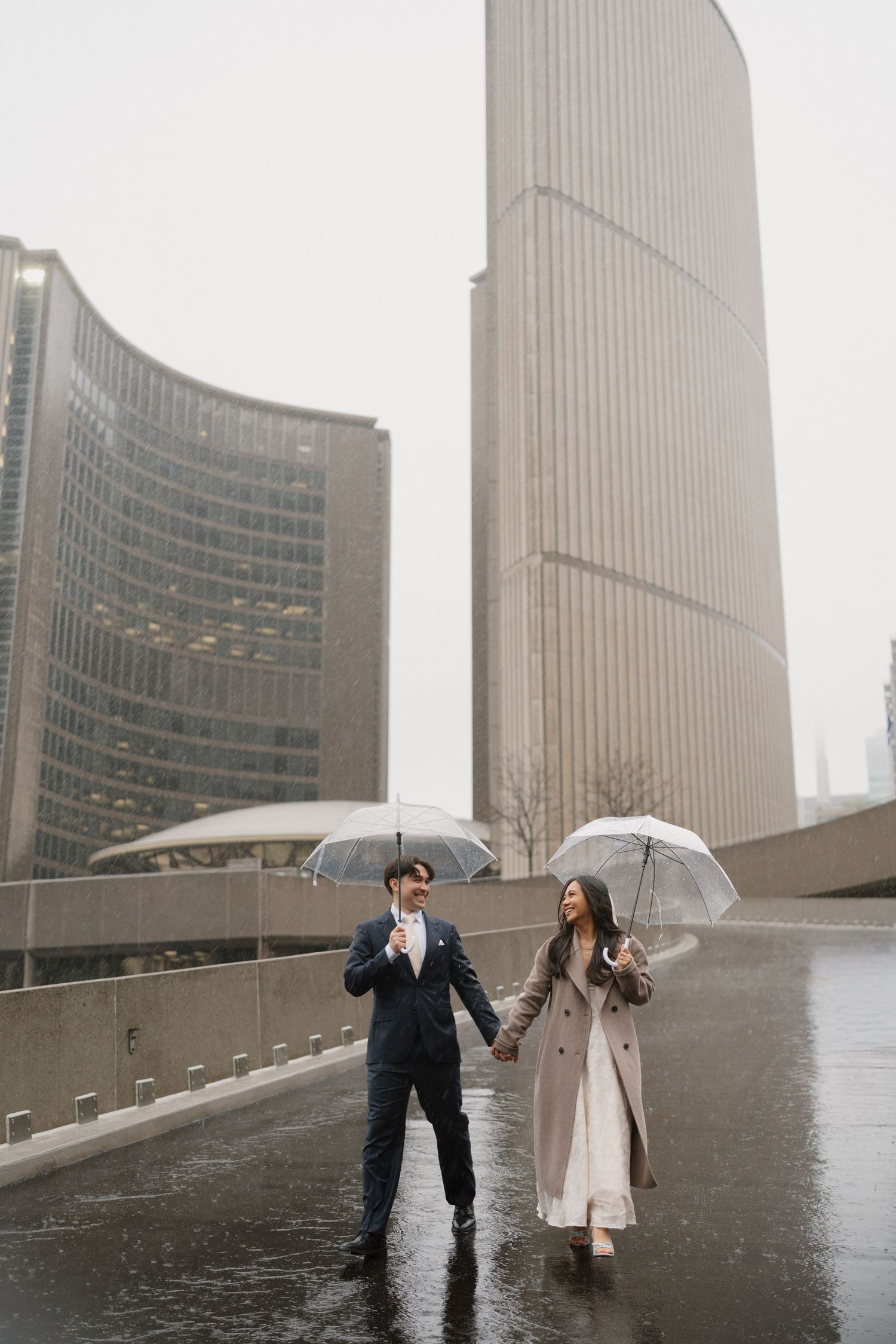Bride and groom hold clear umbrellas while walking hand in hand in the rain with city skyscrapers behind them.