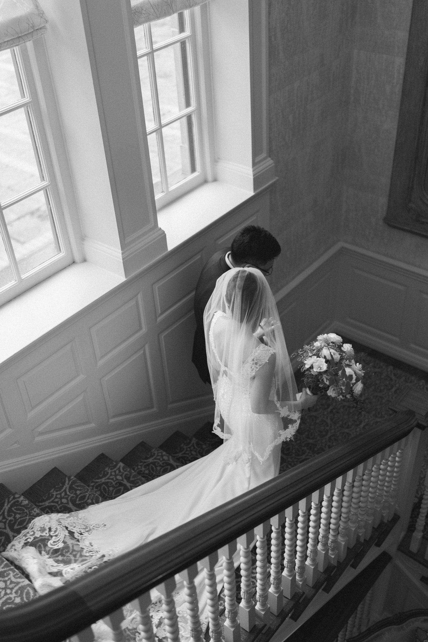 Black and white aerial view of bride and groom walking down grand staircase, bride holding bouquet.