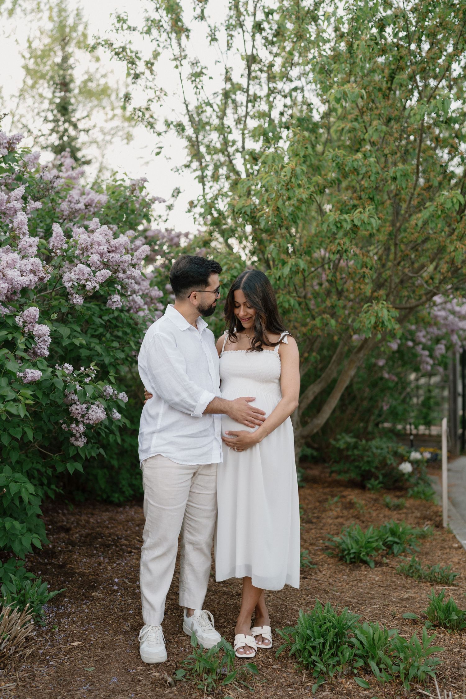 Couple in matching white outfits sharing tender maternity moment beside blooming lilac bushes in garden.