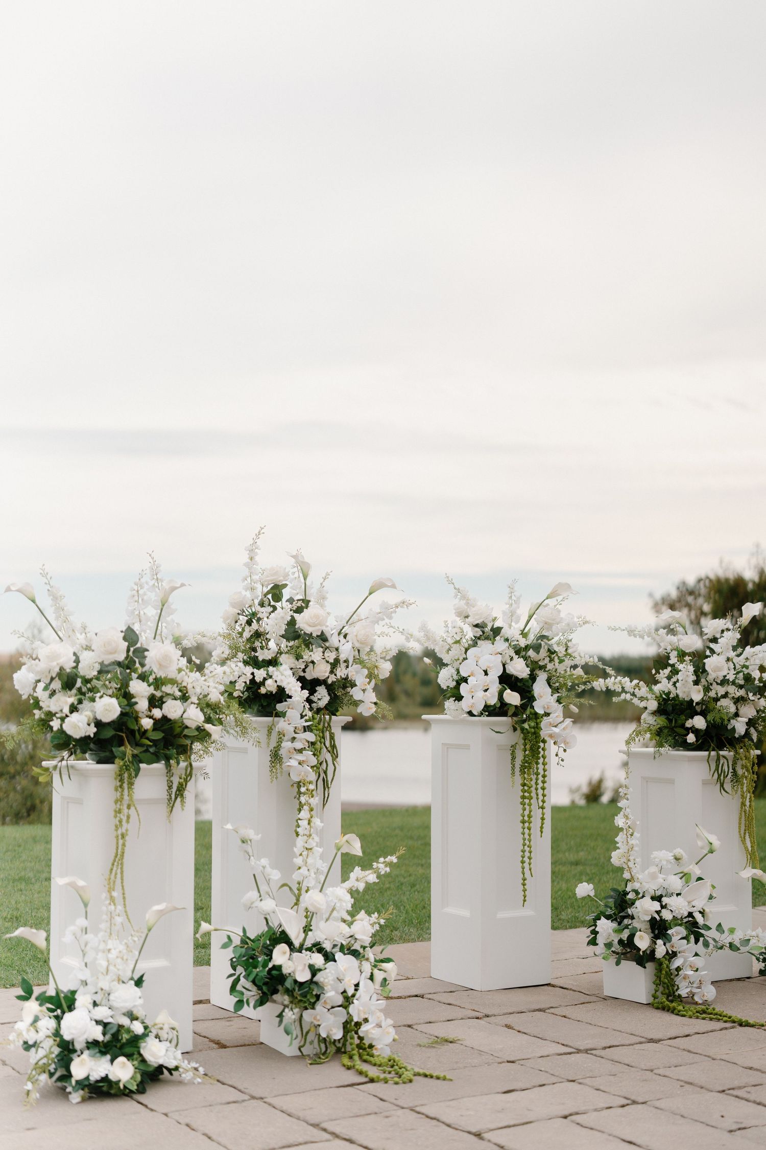 White floral arrangements on tall pedestals at an outdoor wedding ceremony with a serene waterfront backdrop.