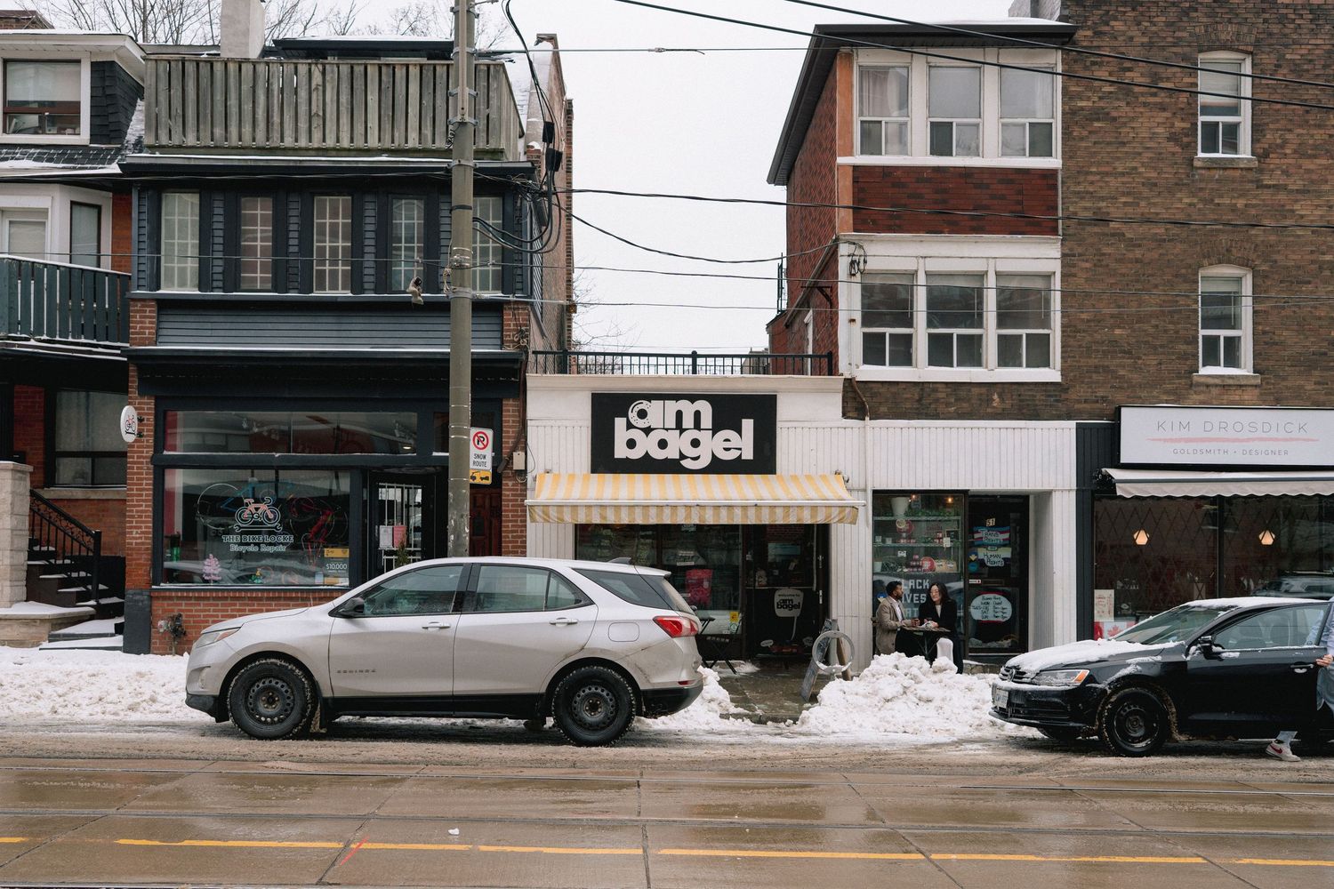 A snowy winter street scene with parked cars and brick buildings along a small town commercial district.