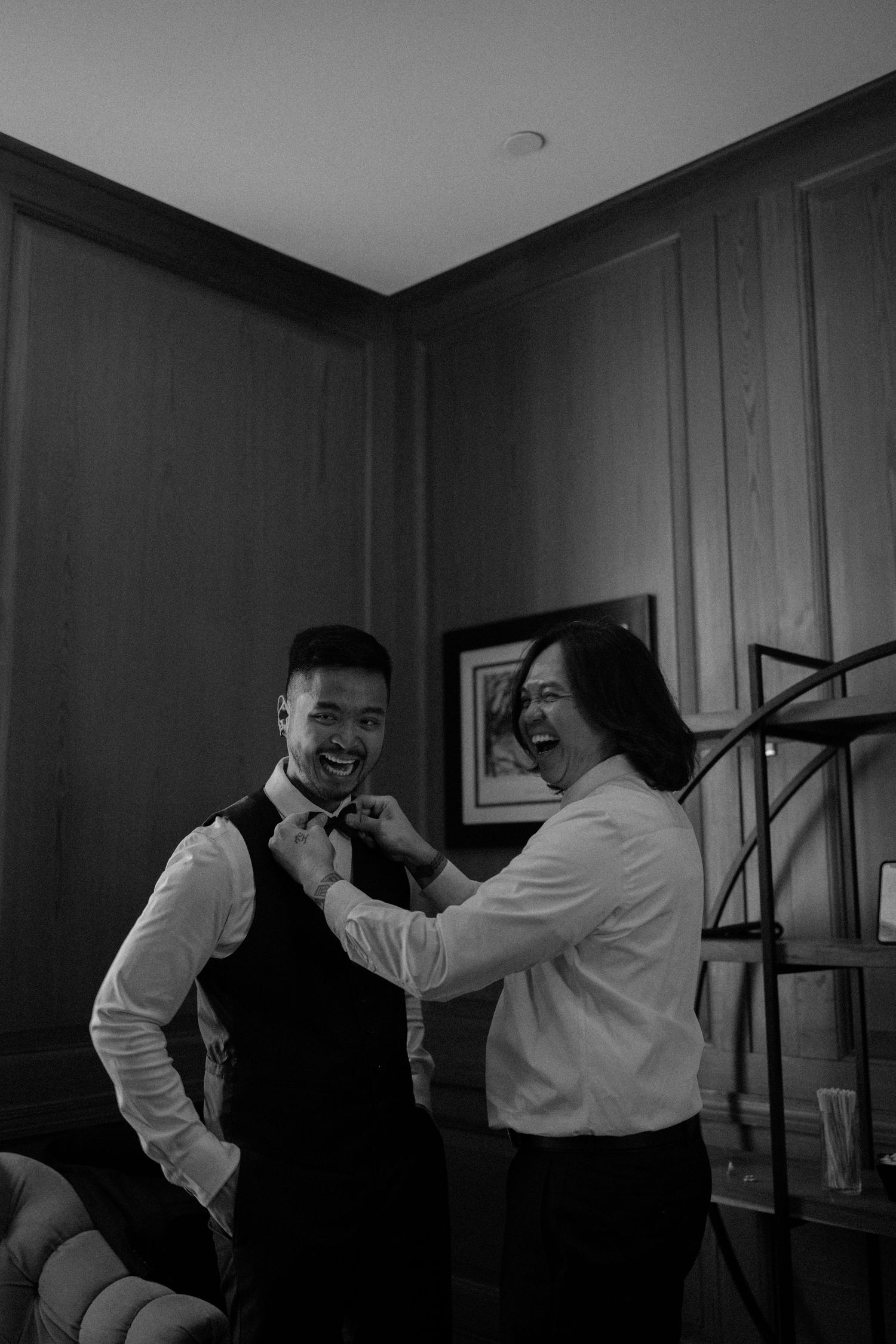 Black and white photo of a formal pre-wedding preparation scene showing bow tie adjustment in elegant indoor setting.