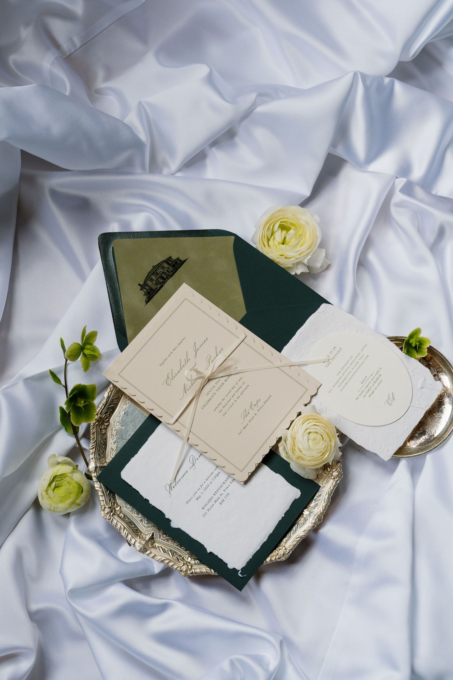 Elegant wedding invitation suite with green envelopes and cream cards displayed on satin with white roses.