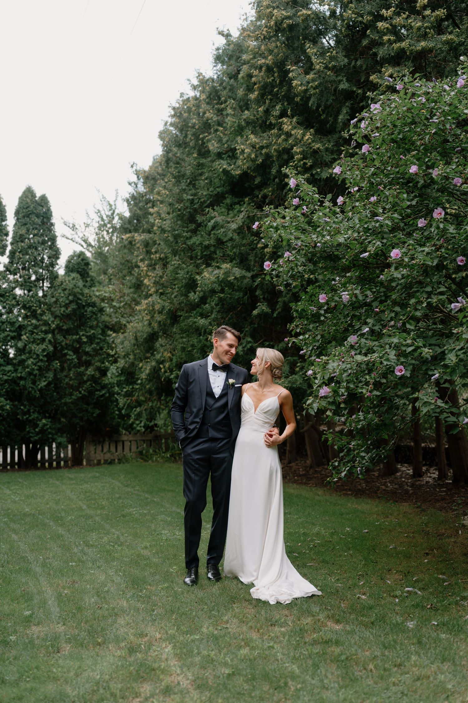 Bride and groom standing together on green lawn surrounded by lush trees with purple blooms on their wedding day.