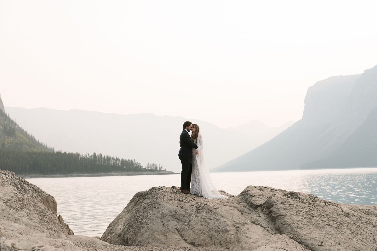 Bride and groom share a kiss on rocky shoreline with misty mountain lake backdrop.
