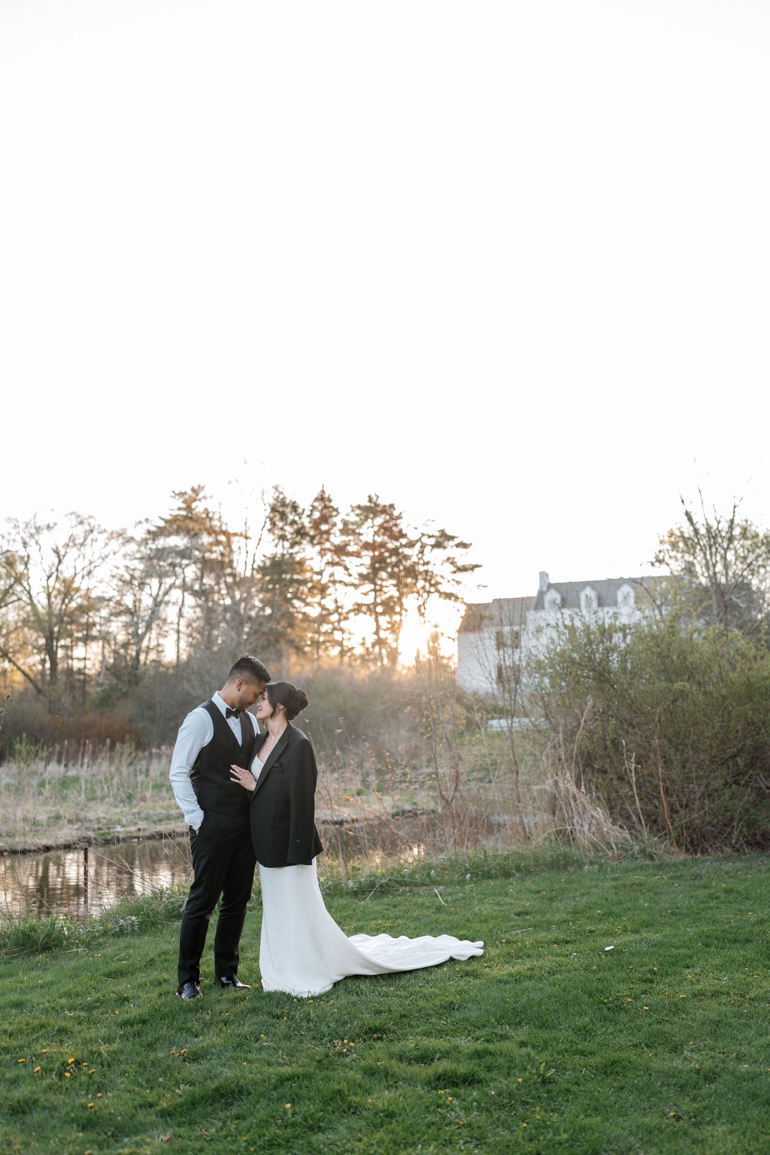 A couple shares an intimate moment at sunset on a grassy lawn with trees and a pond in the background.