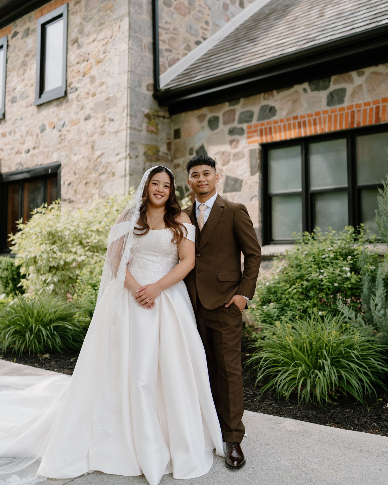 Bride in white gown and groom in brown suit pose together outside a stone building on their wedding day.
