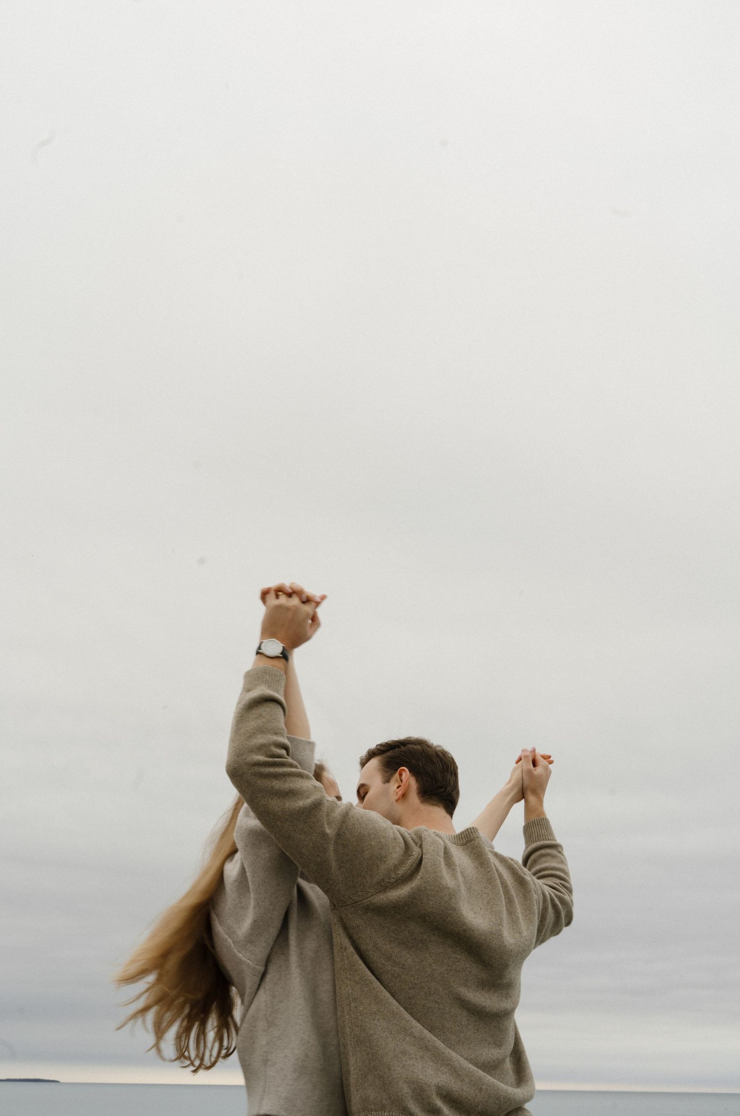 Couple holding hands raised against overcast sky, wearing matching gray sweaters, woman's hair blowing in wind.