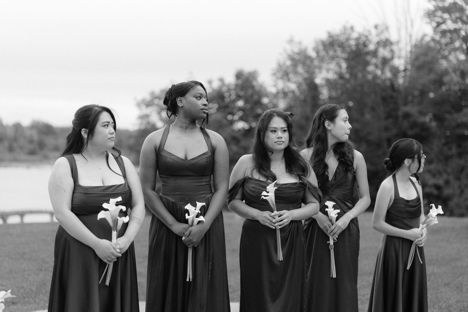 Bridesmaids in matching dark gowns holding white flowers stand outdoors near a lake in a black and white wedding photo.