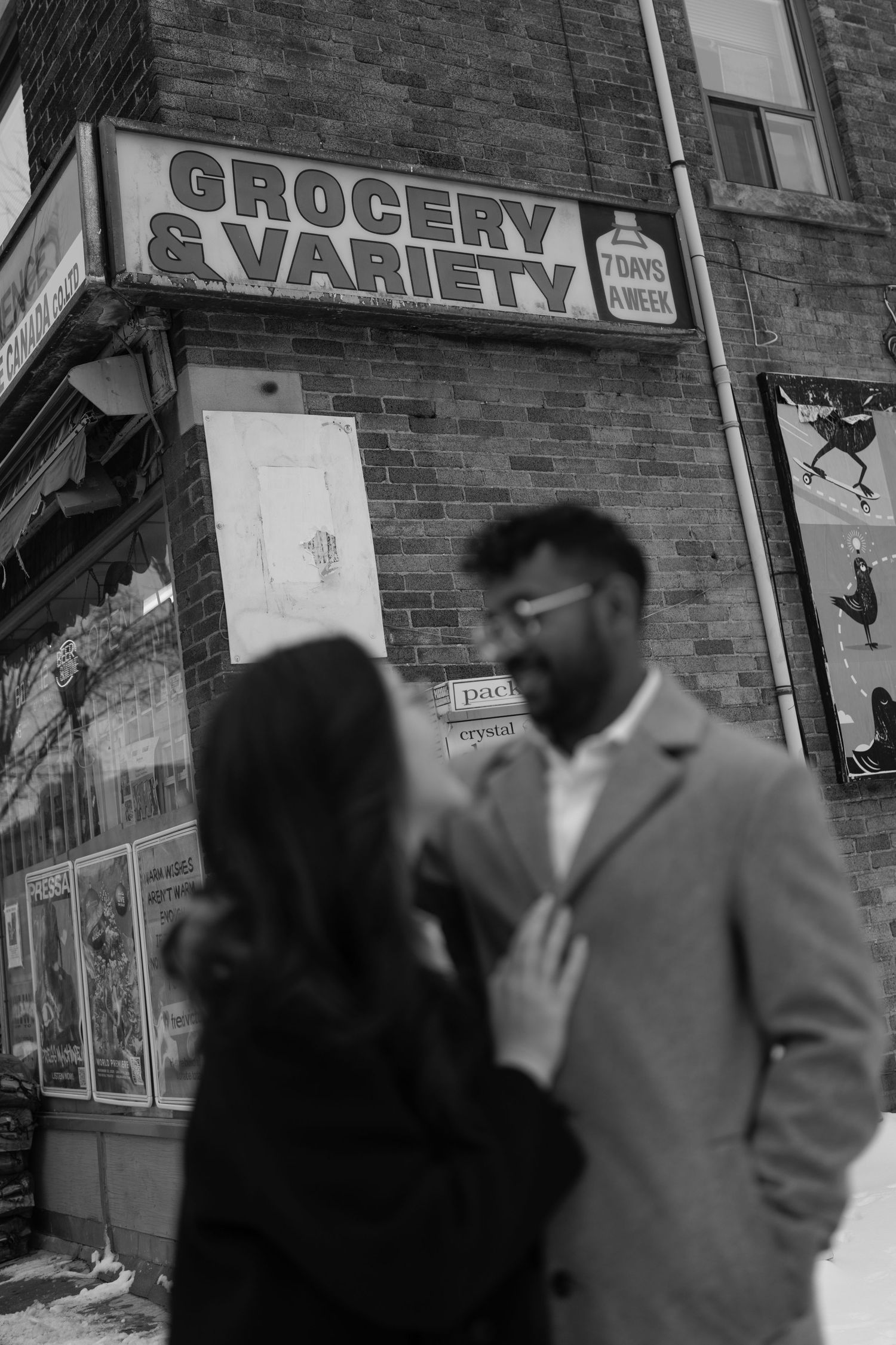Black and white photo of a couple walking past storefronts with grocery and variety shop signs visible.