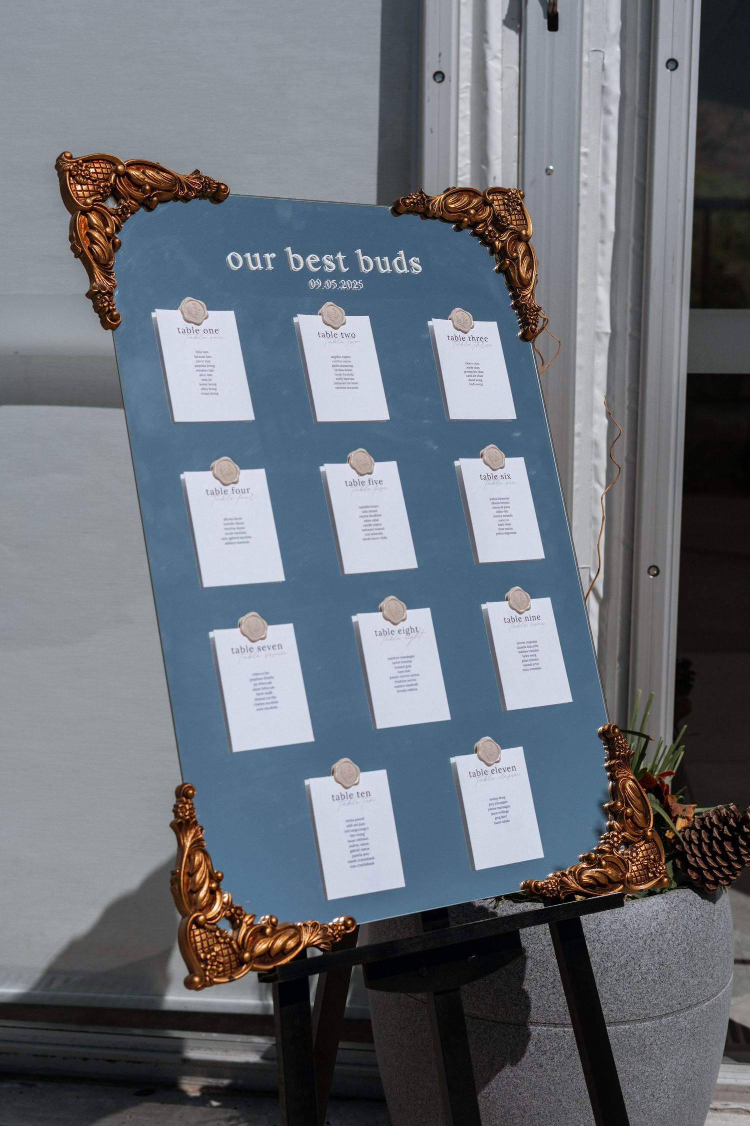 A blue wedding seating chart display with ornate gold corner decorations shows paper cards arranged in grid formation.