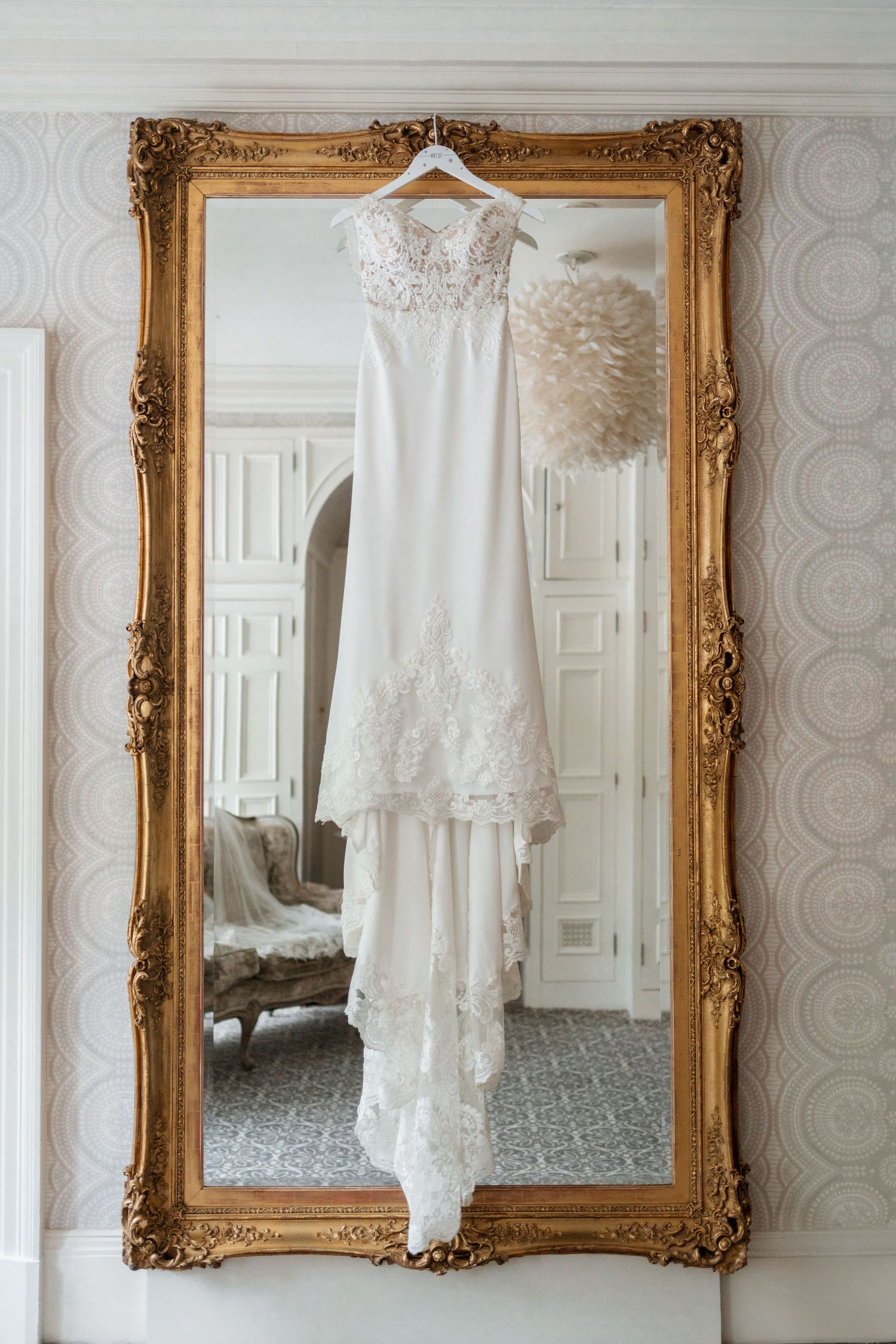 Elegant white lace wedding dress hanging from gold ornate mirror in luxurious room with patterned wallpaper.