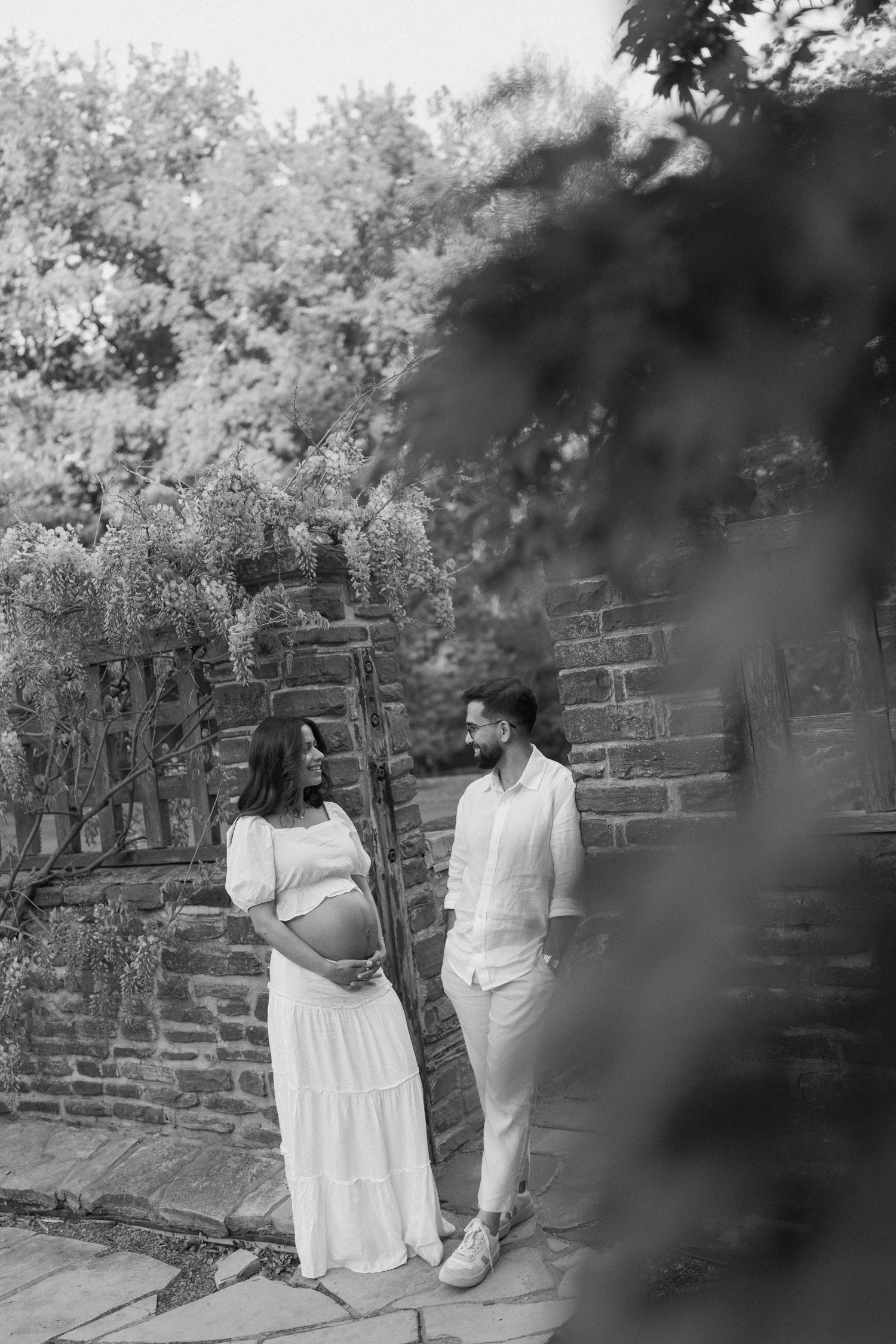 Black and white maternity photo of couple in white outfits standing by stone wall with wisteria in lush garden.
