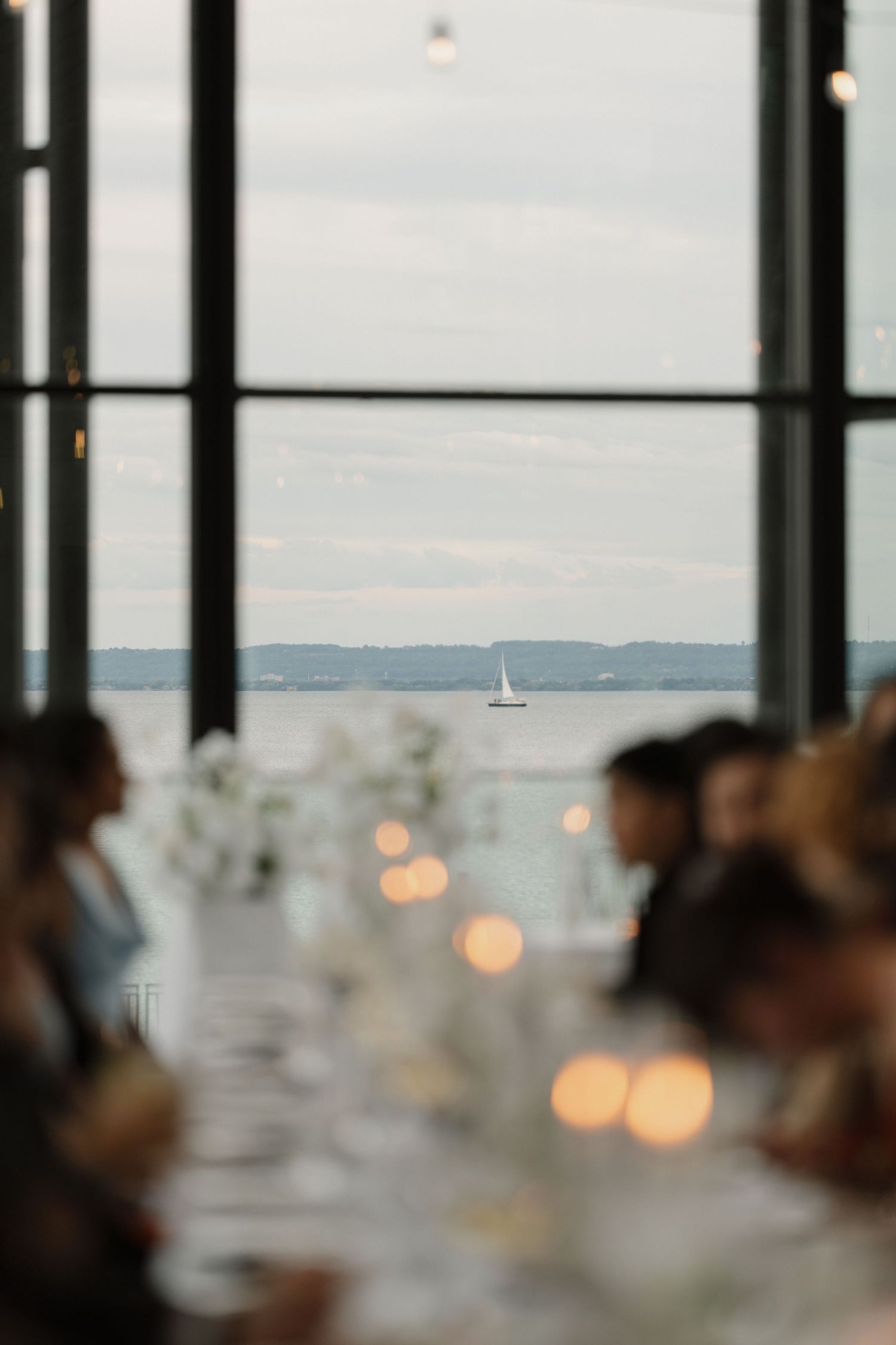 Elegant waterfront dinner reception with candlelit tables and guests, sailboat visible through large windows over calm sea.