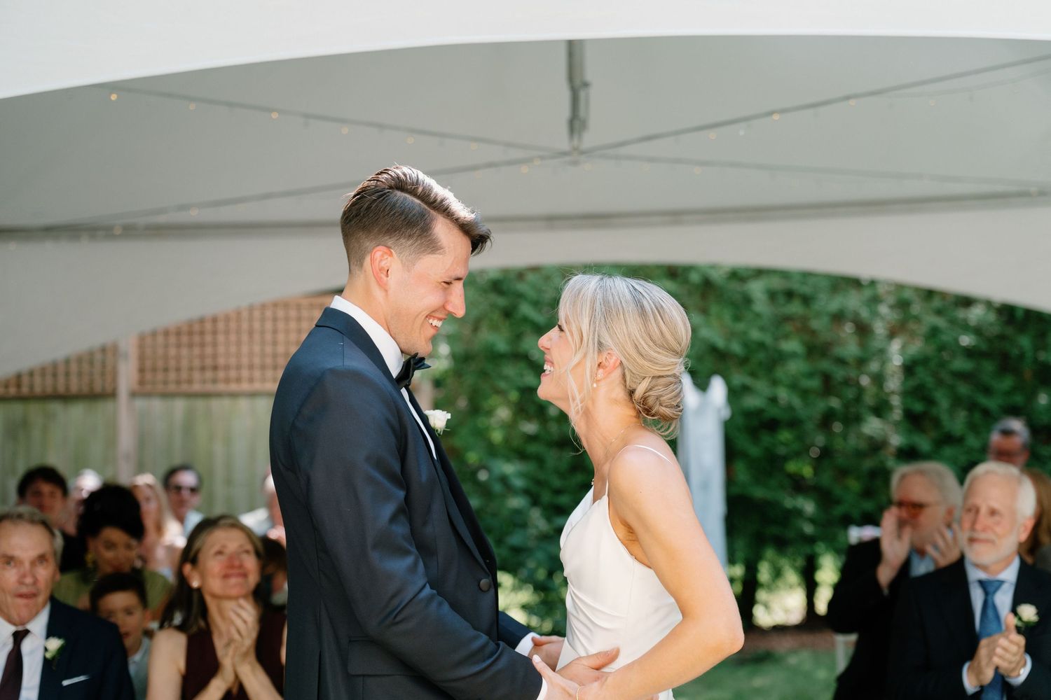 Bride and groom share a joyful moment during outdoor wedding ceremony under white tent with guests applauding.