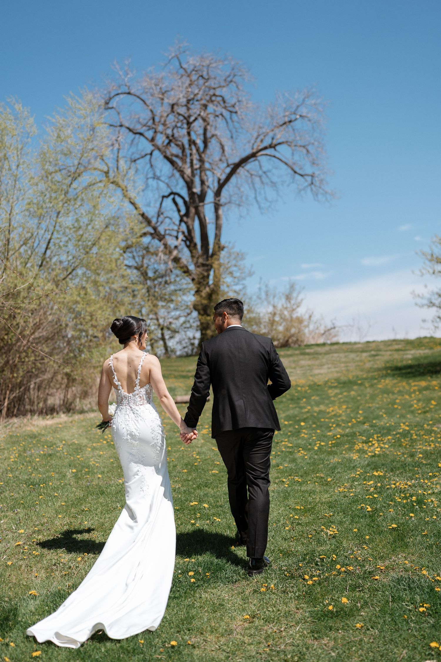 A couple in wedding attire walks together on a grassy hill dotted with yellow dandelions under a bright blue spring sky.