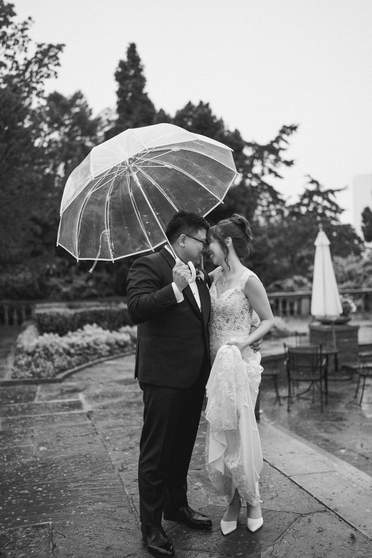 Black and white photo of newlyweds sharing an intimate moment under a clear umbrella on a rainy patio.