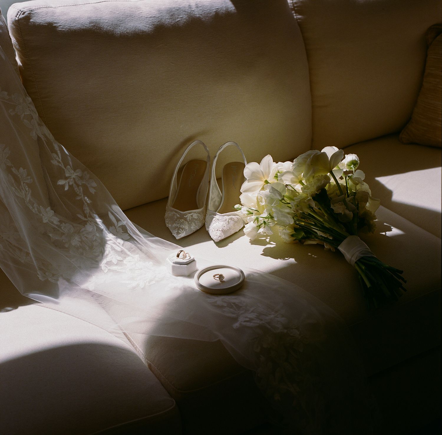 Wedding details with white bouquet, bridal shoes and ring on beige couch in natural sunlight.