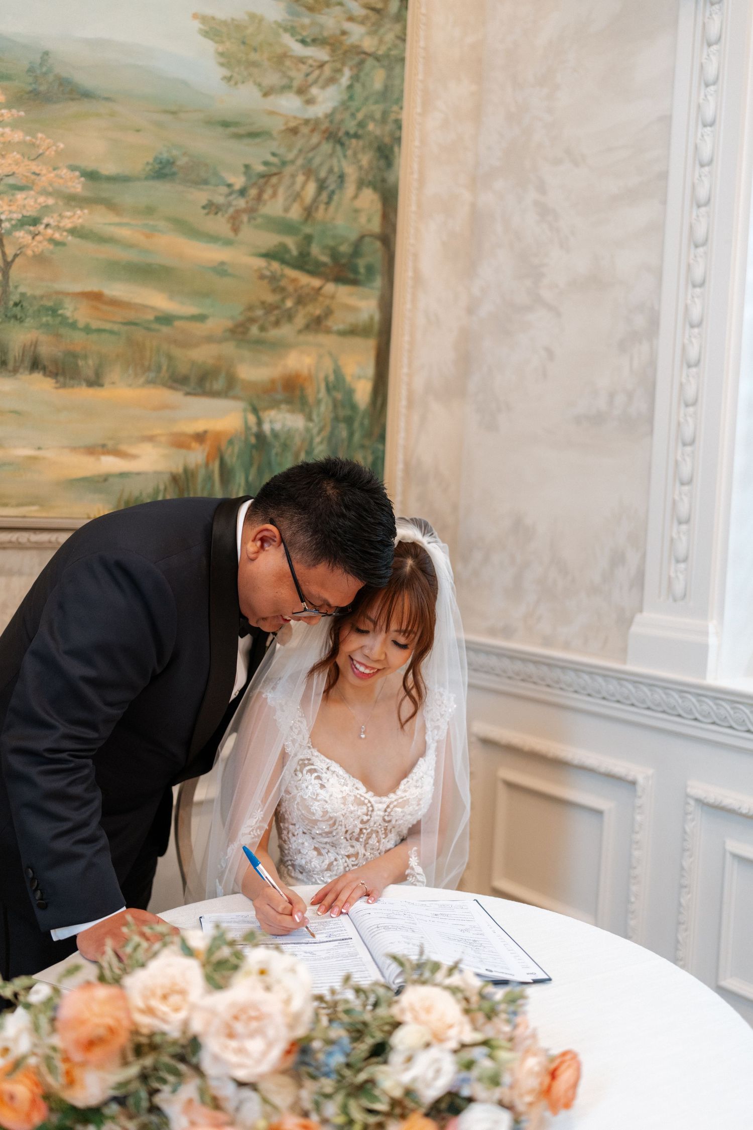 A couple signs marriage documents at an elegant venue with peach floral arrangements on the table.