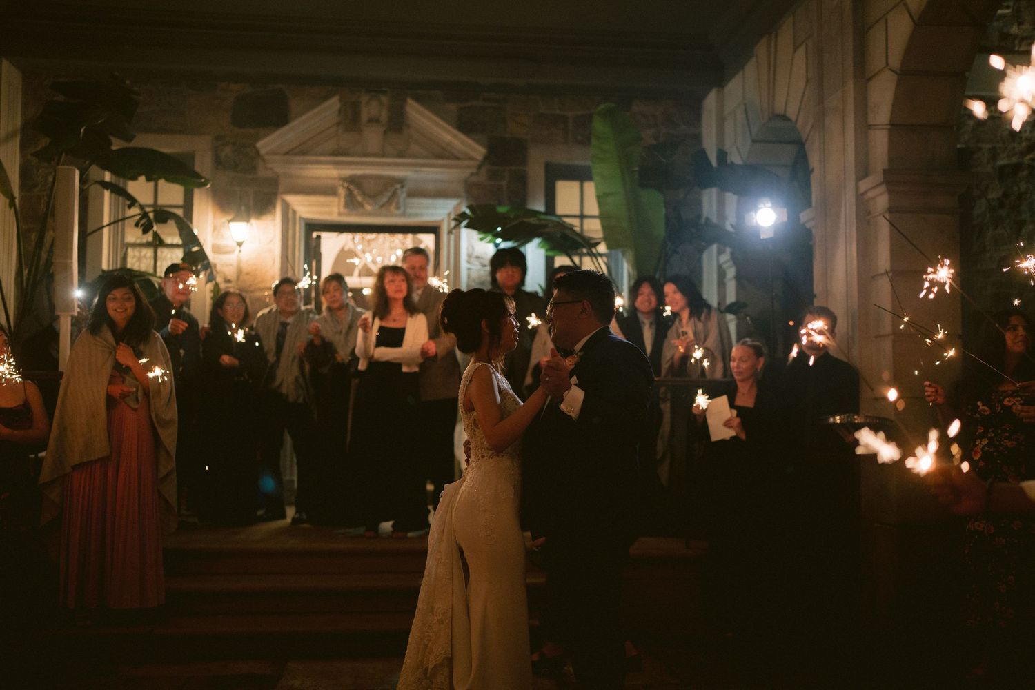 Bride and groom exit wedding reception through sparkler send-off with guests lining the path at night.