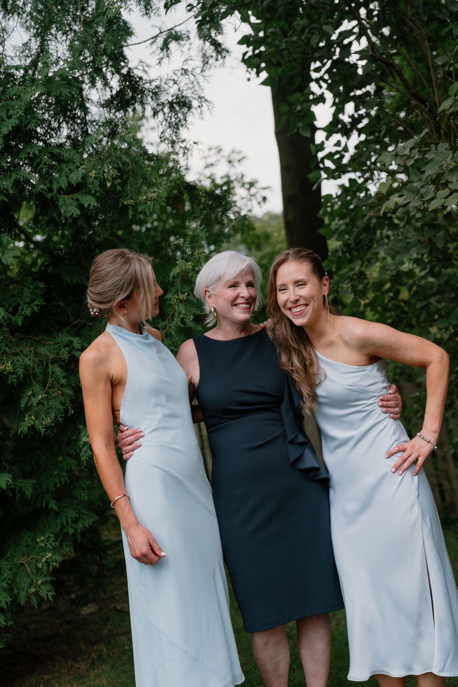 Three women in elegant dresses smiling together outdoors among lush green trees at a wedding celebration.