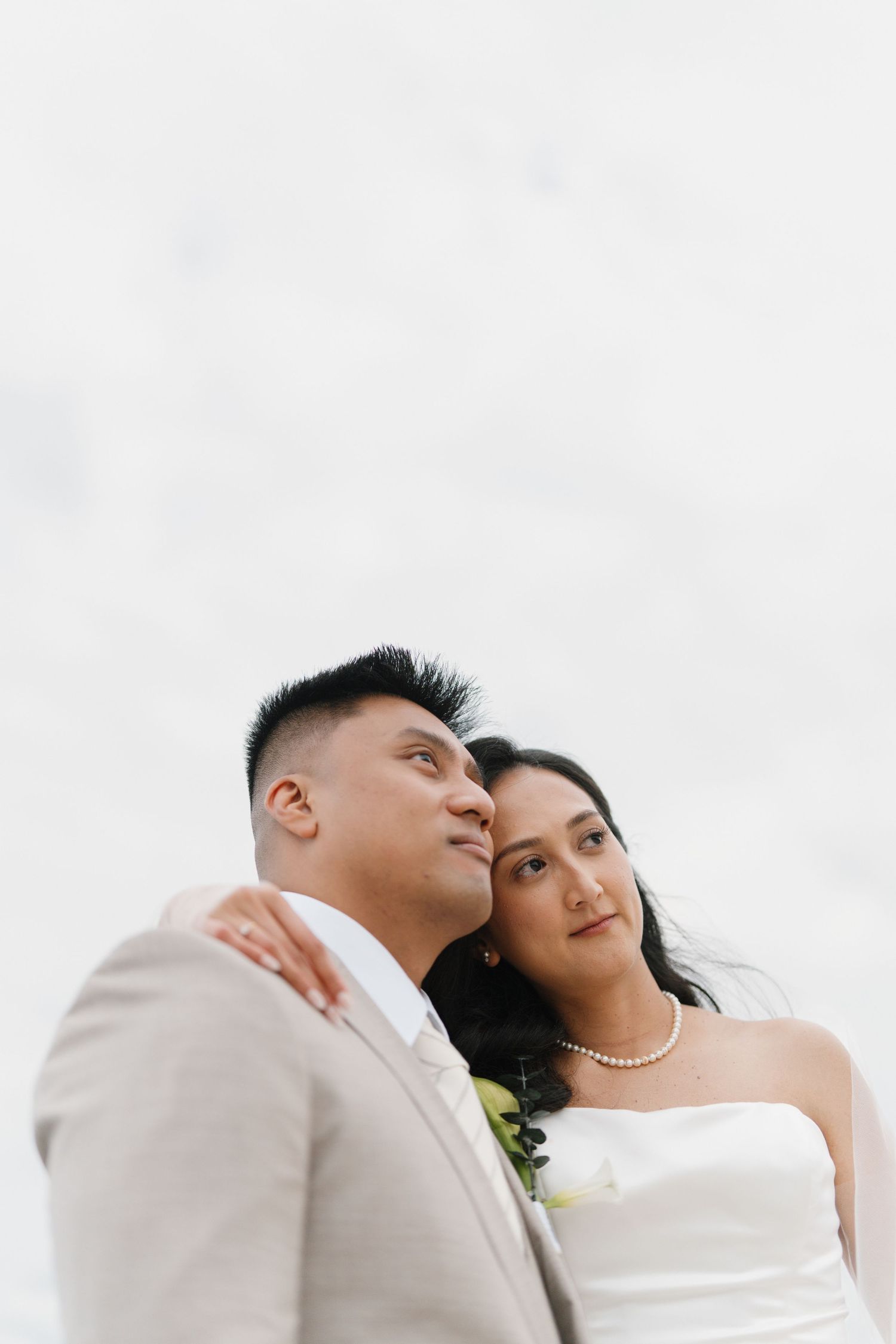 A bride in a white strapless gown and groom in a beige suit pose together outdoors on their wedding day.
