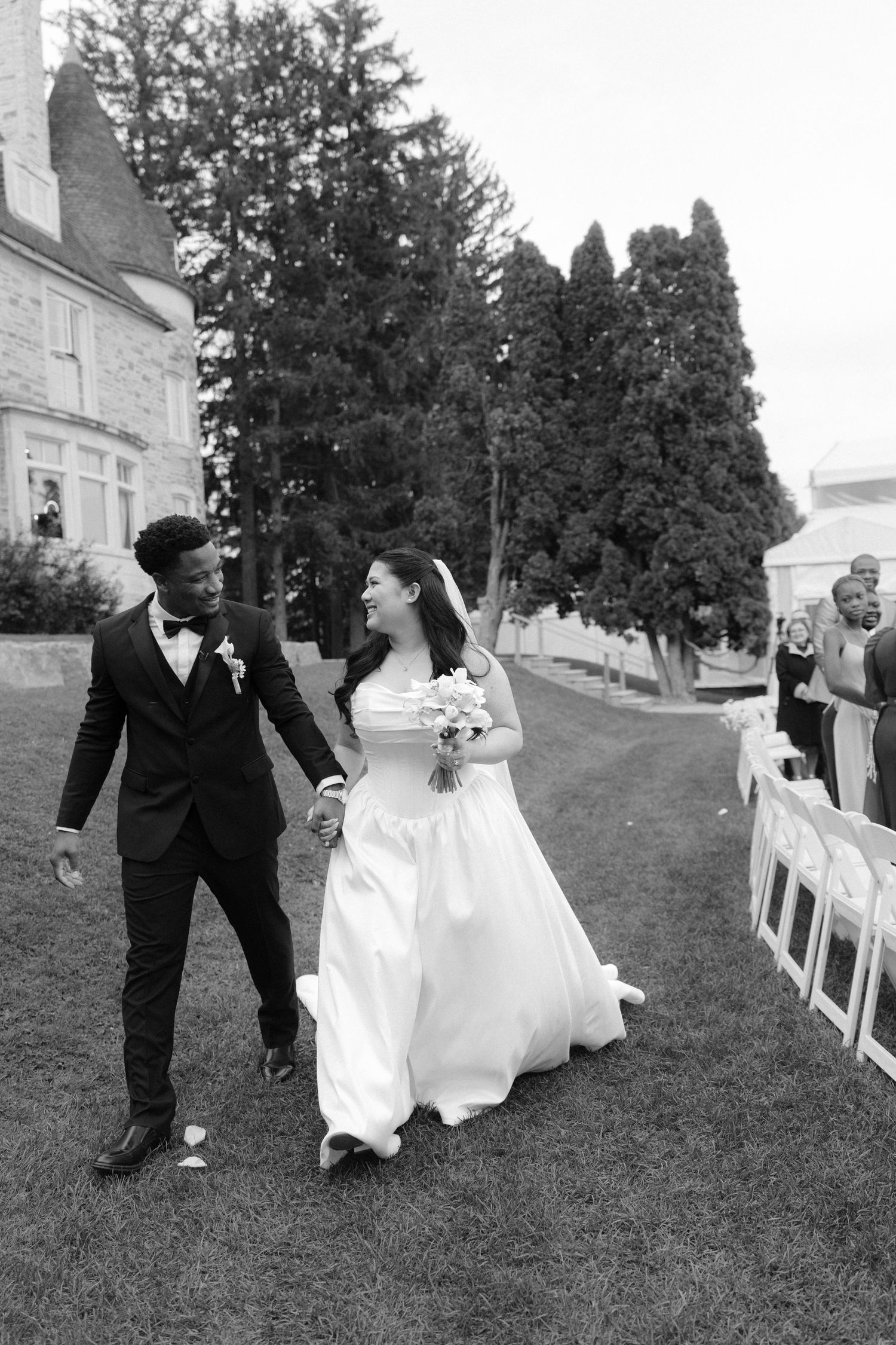 Black and white photo of a smiling interracial couple walking hand in hand down the aisle after their outdoor wedding ceremony.