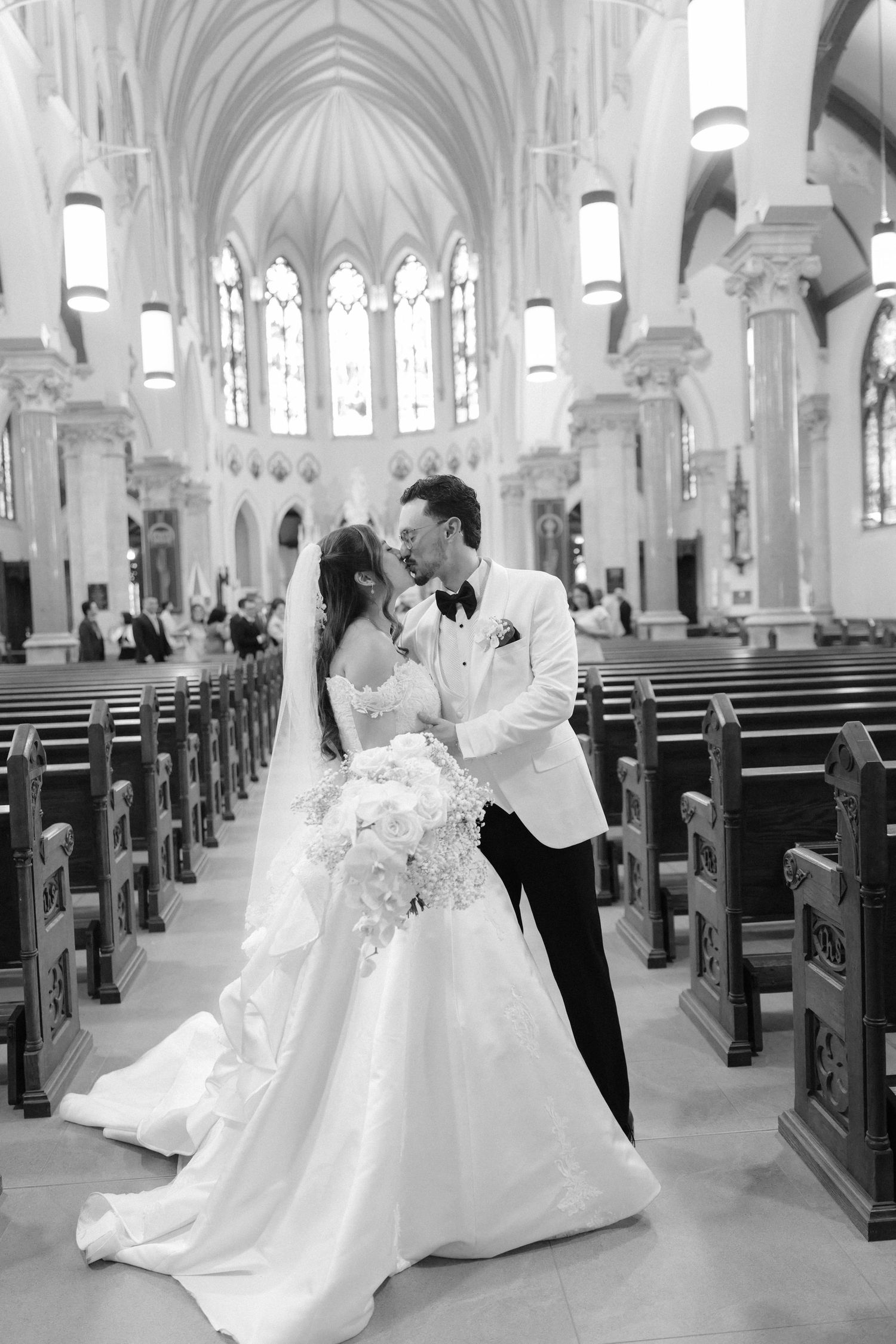 Black and white photo of a bride and groom sharing a kiss in a grand church aisle, bride holding a bouquet.