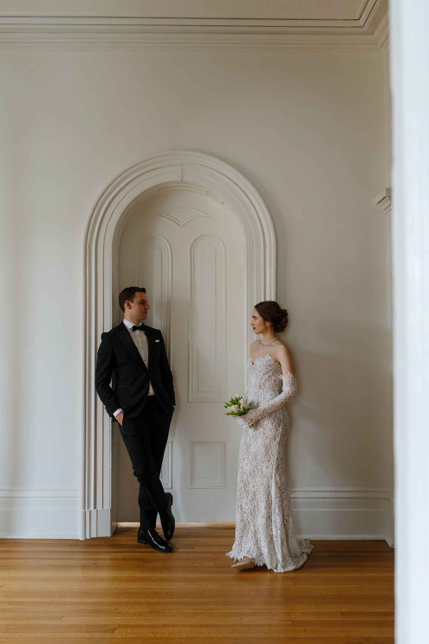 A bride in lace gown and groom in black tuxedo stand facing each other in an elegant white arched hallway.