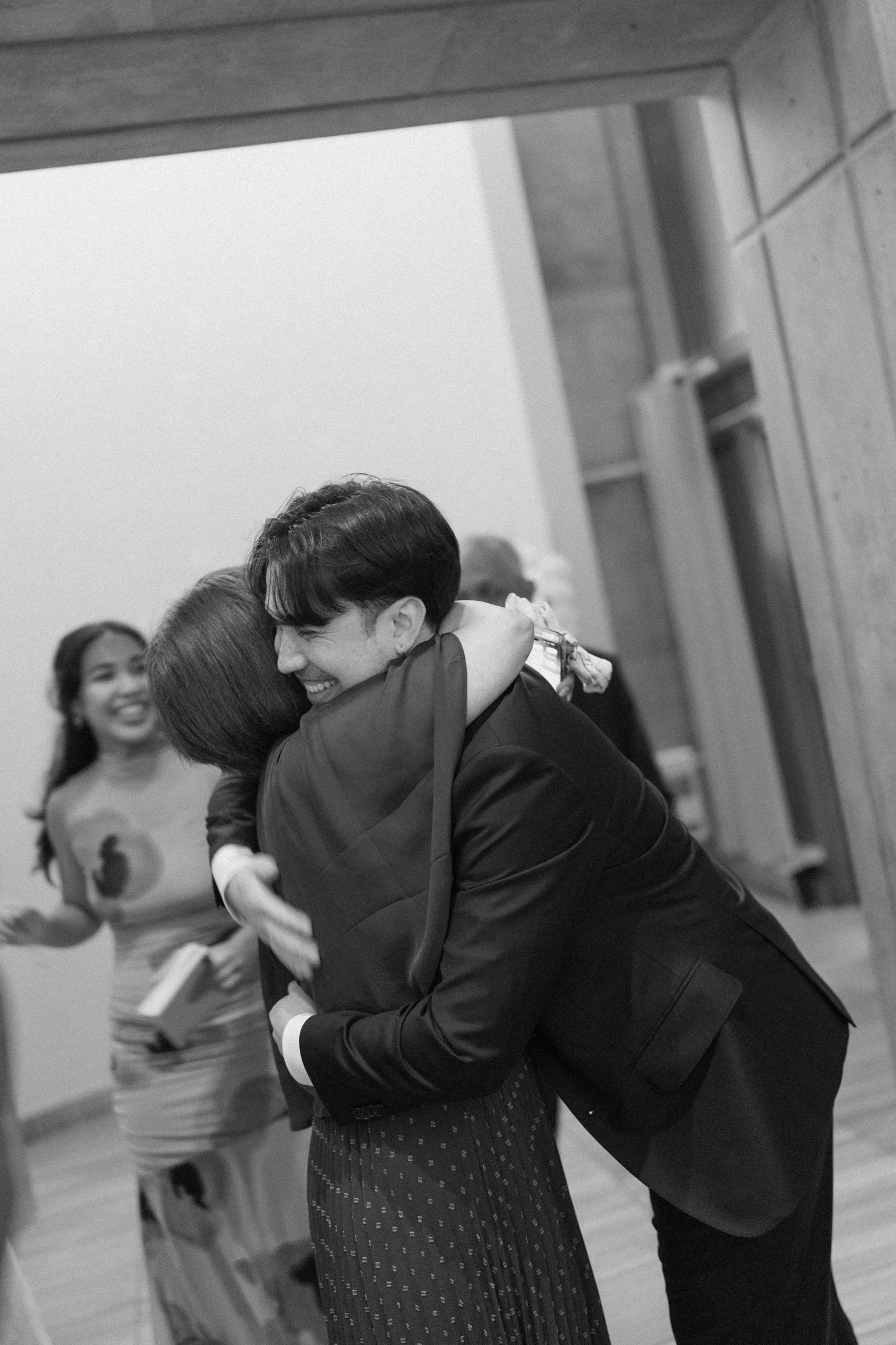 Black and white photo of two people sharing a warm embrace at a formal event, with smiling guests in the background.