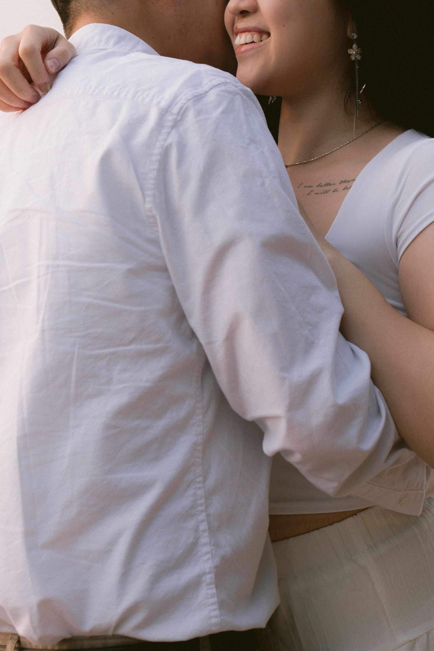 Couple embracing in matching white outfits, woman smiling with a tattoo visible on her chest.