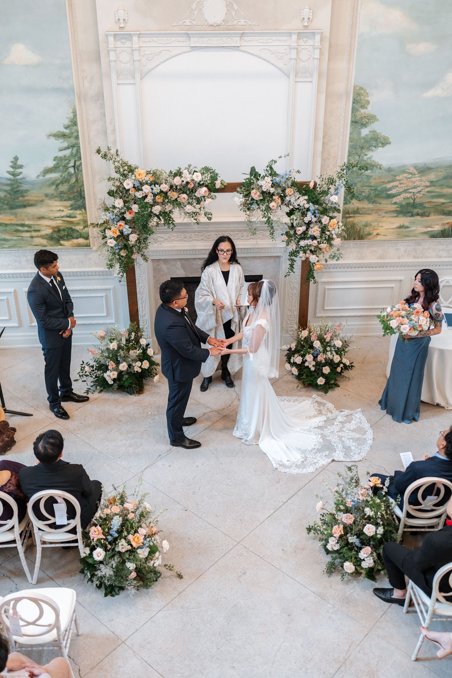 Indoor wedding ceremony featuring elegant floral arch arrangements and white decor at an ornate venue with painted landscape walls.