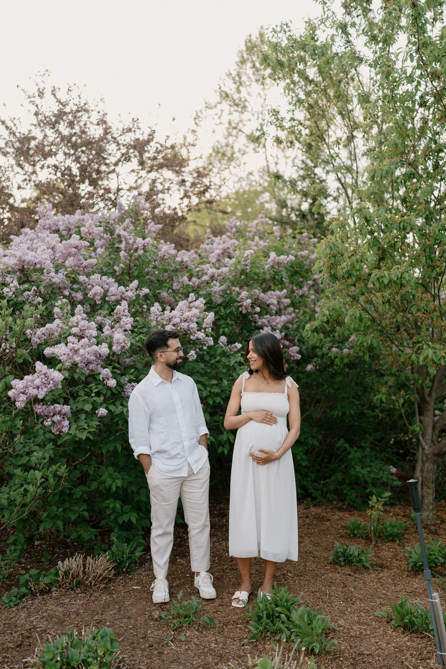 Pregnant couple in white outfits standing together in a garden with blooming lilac bushes during golden hour.