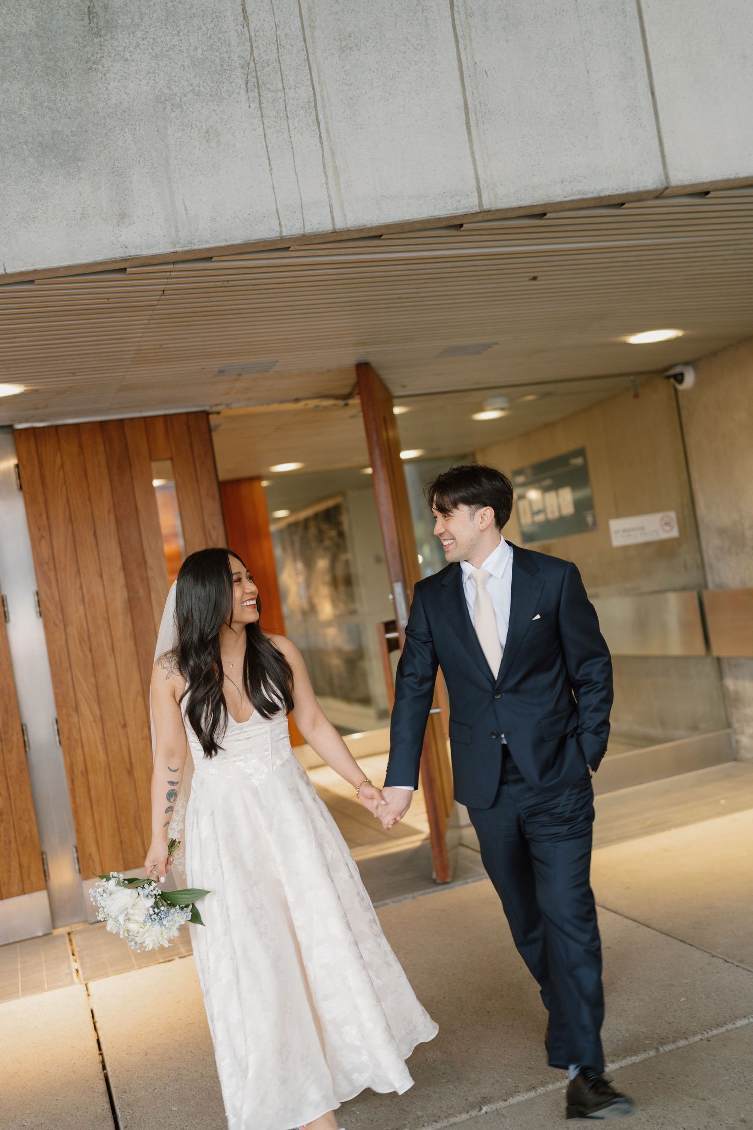 Bride and groom walk hand in hand outside a modern building, she holds white flowers in a white gown, he wears a dark suit.