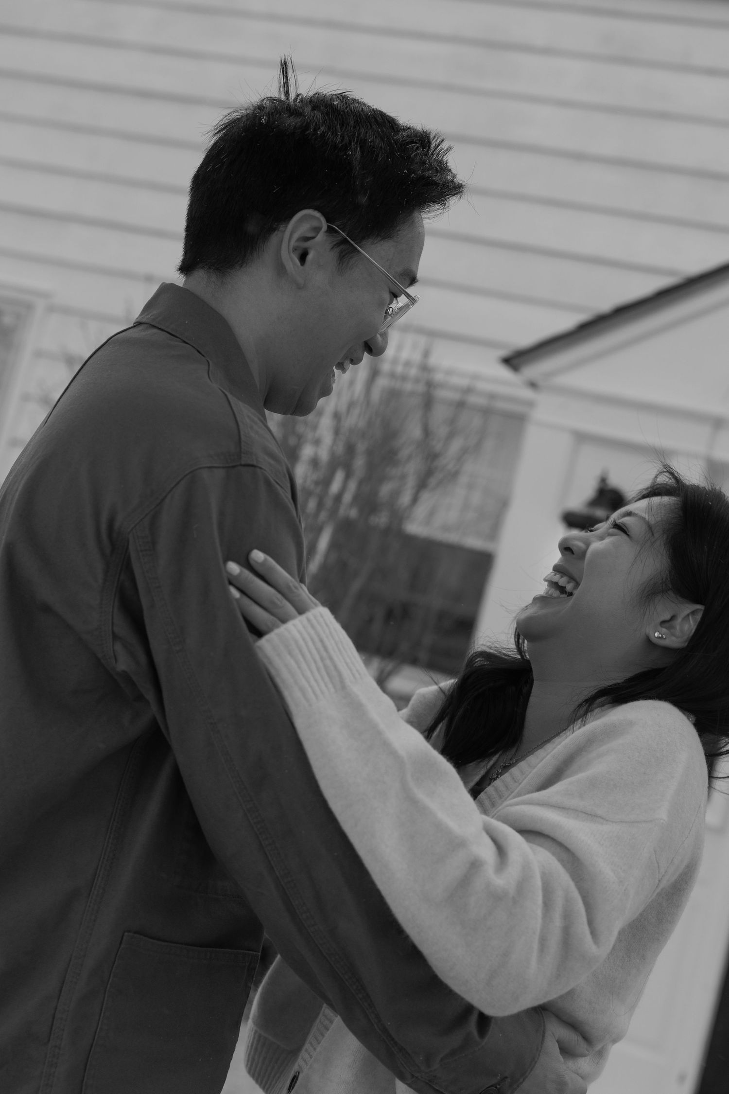 Black and white photo of laughing Asian couple embracing and gazing at each other outdoors.