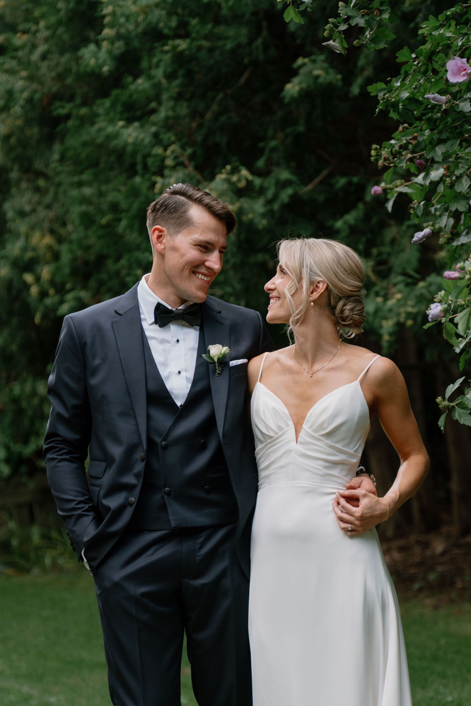 A smiling bride and groom in formal wedding attire stand together outdoors among lush green trees and blooming flowers.