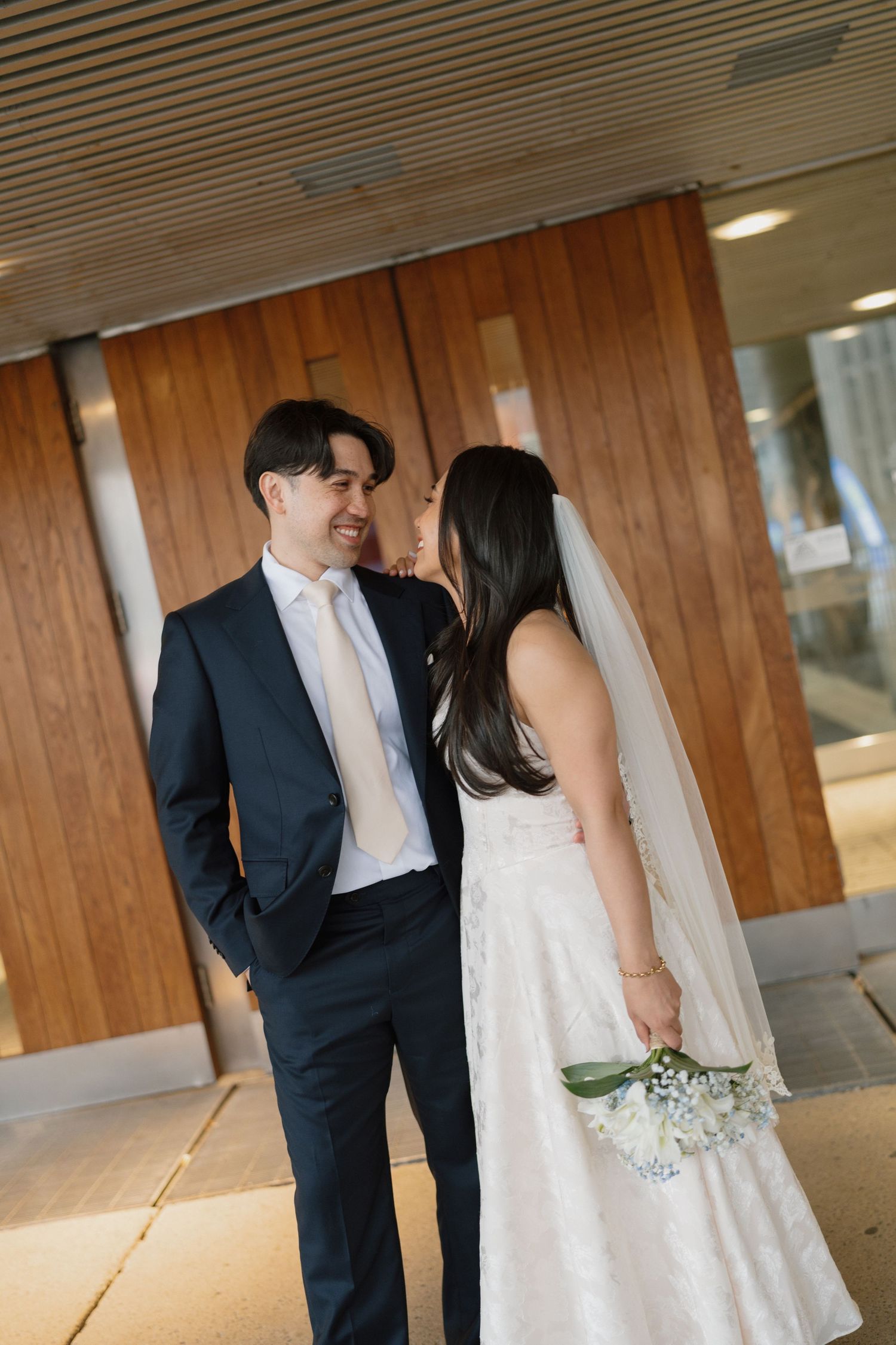 Bride and groom share a romantic moment, she holds a white bouquet in a lace dress with veil, he wears a navy suit.
