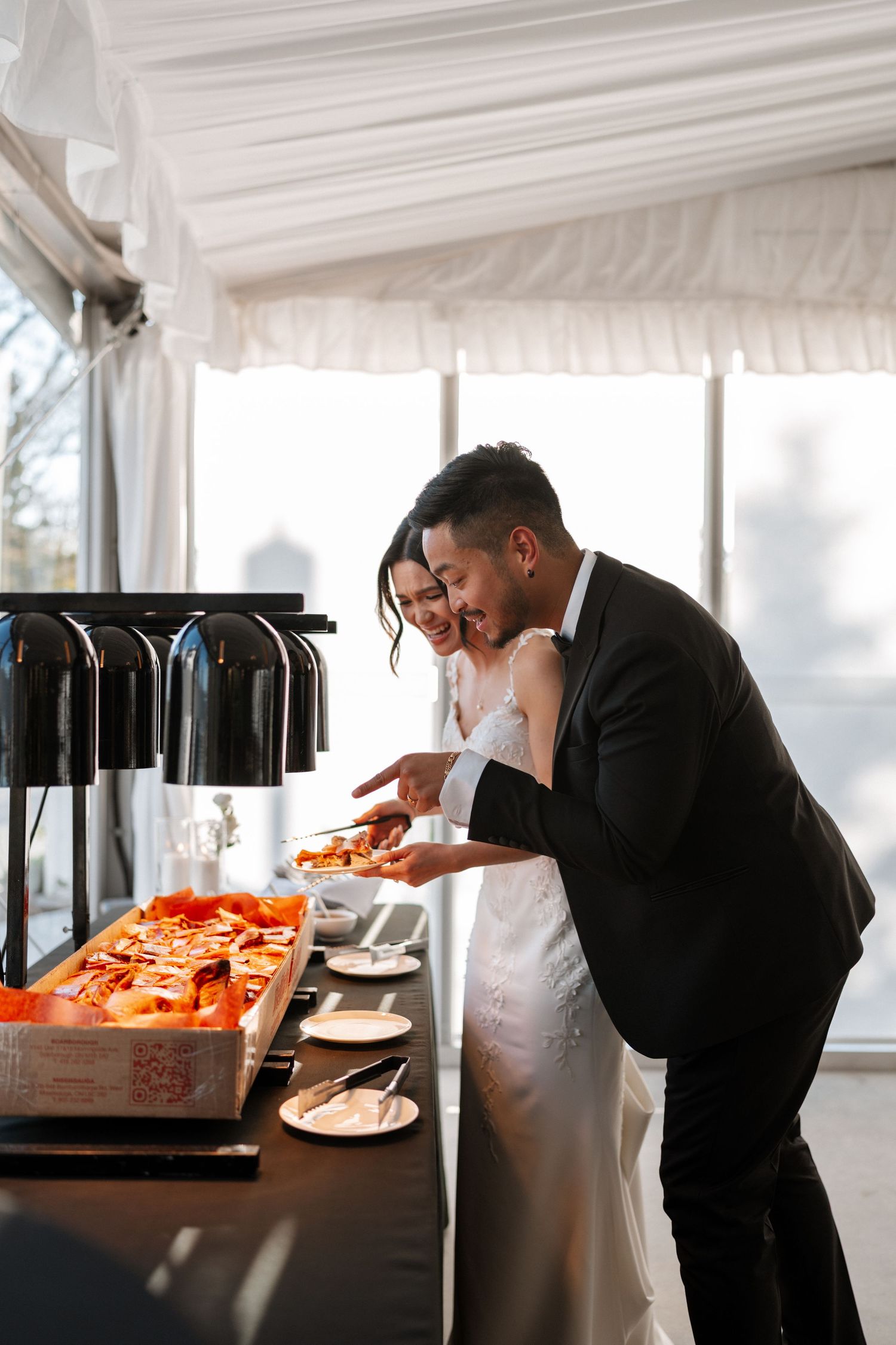 A couple at a wedding reception buffet smiling while serving themselves food from warming dishes in an elegant white tent.