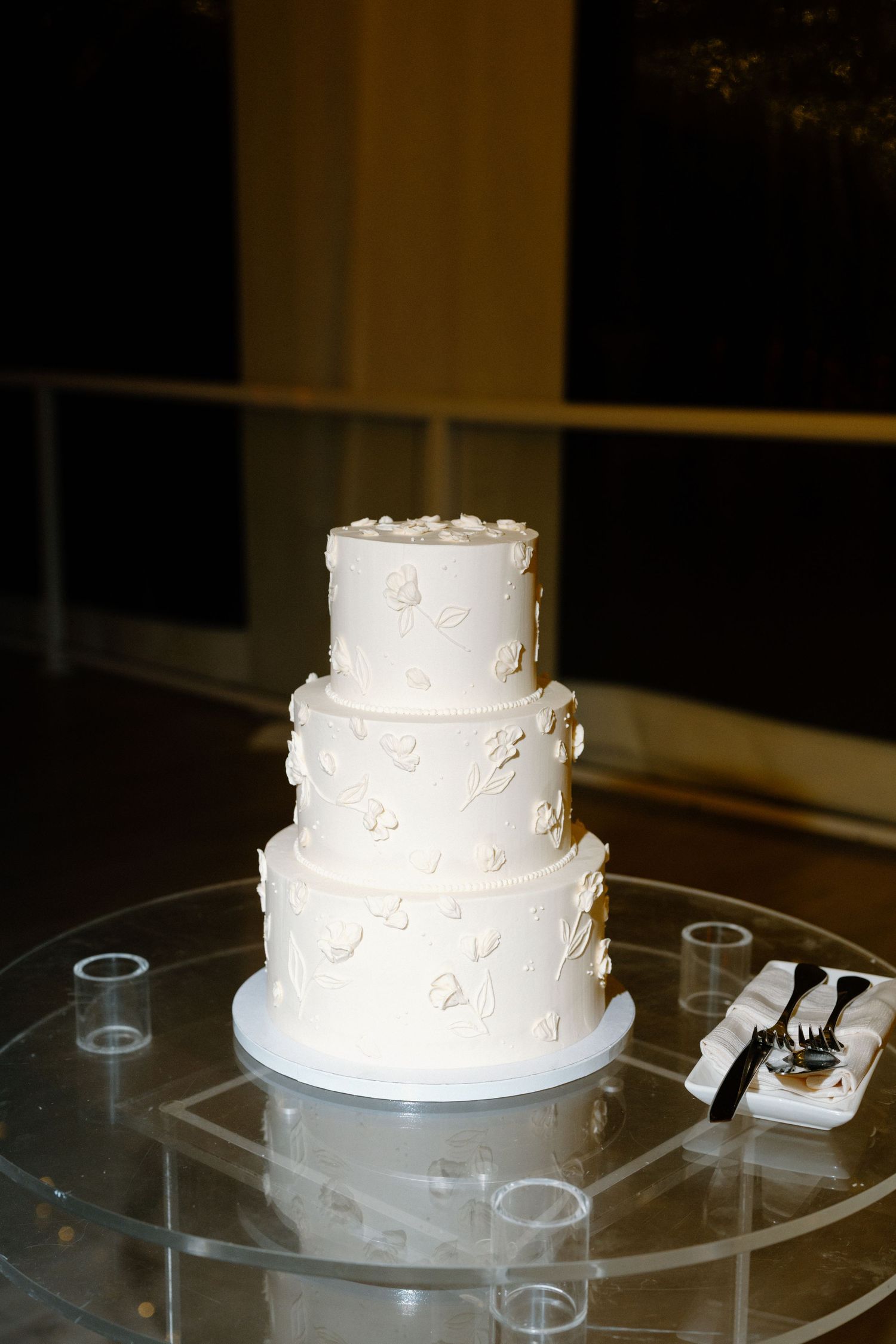 Elegant three-tier white wedding cake with floral details on a glass table, set against a dark evening backdrop.