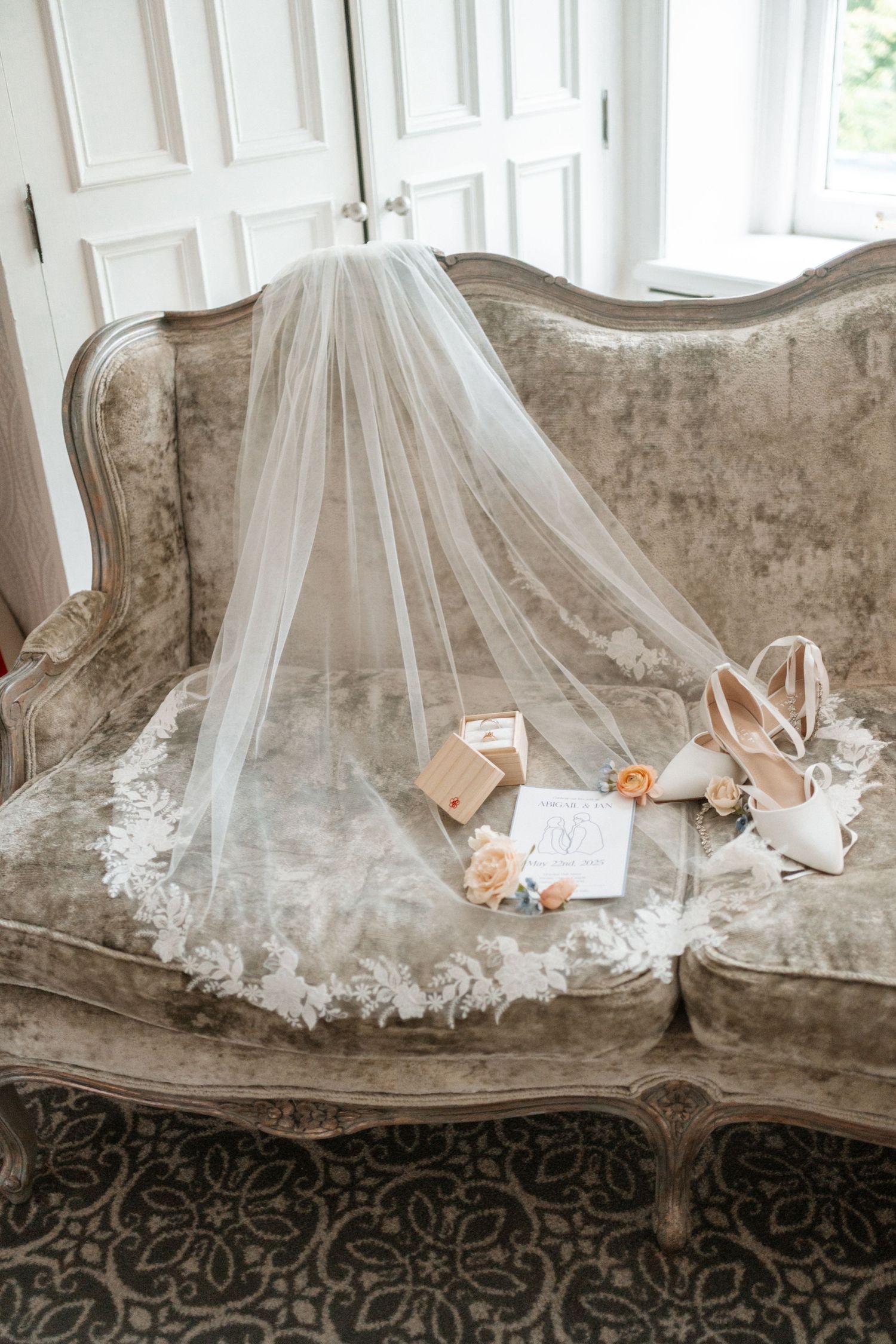 Wedding accessories including veil, shoes and stationery delicately arranged on an elegant vintage velvet sofa.