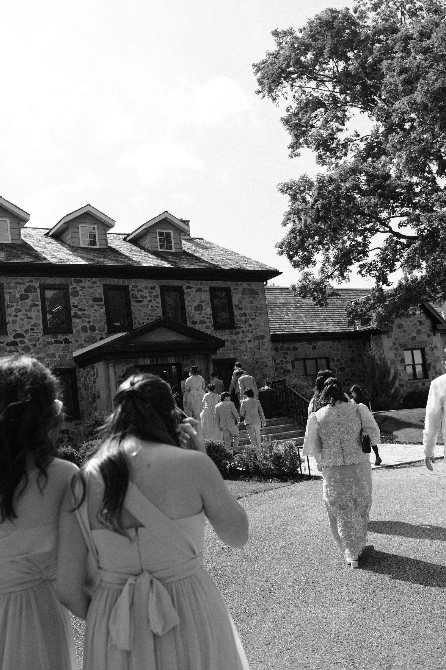Black and white photo of bridesmaids in white dresses walking toward a stone wedding venue with a large tree overhead.