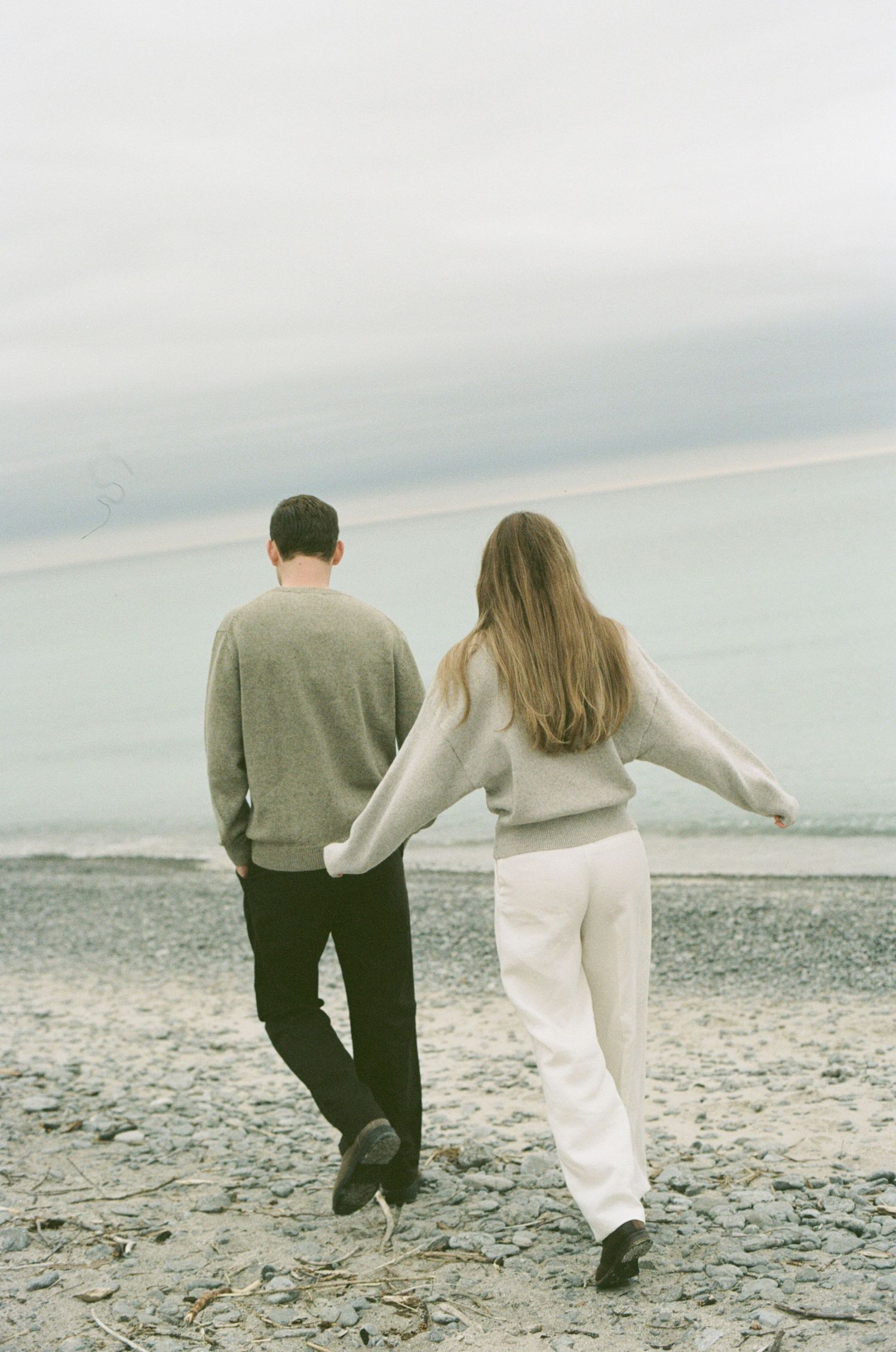 Couple walking hand in hand on a rocky beach, wearing neutral sweaters, viewed from behind with calm ocean horizon.