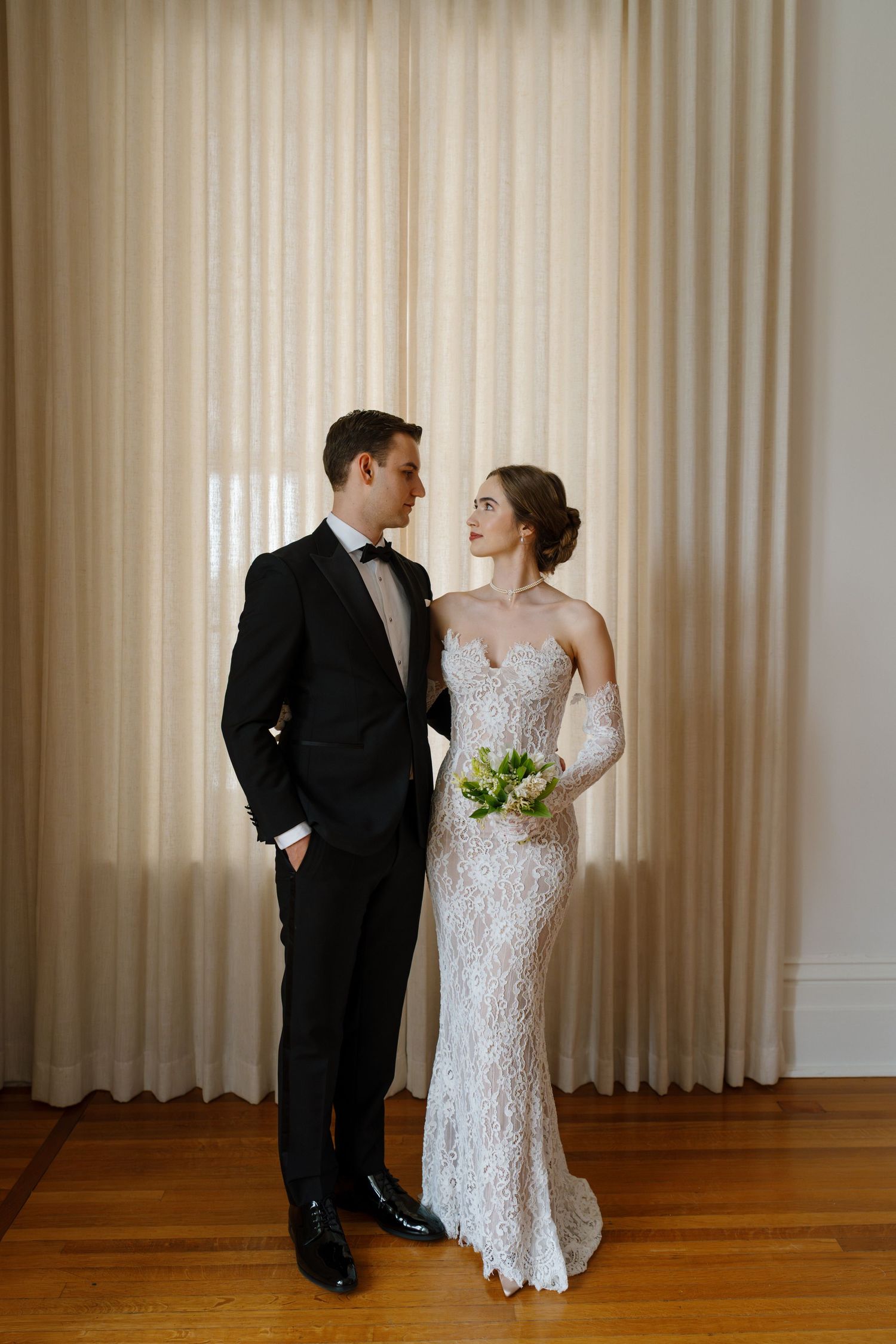 Bride and groom posing together in elegant wedding attire before sheer curtains on hardwood floors.