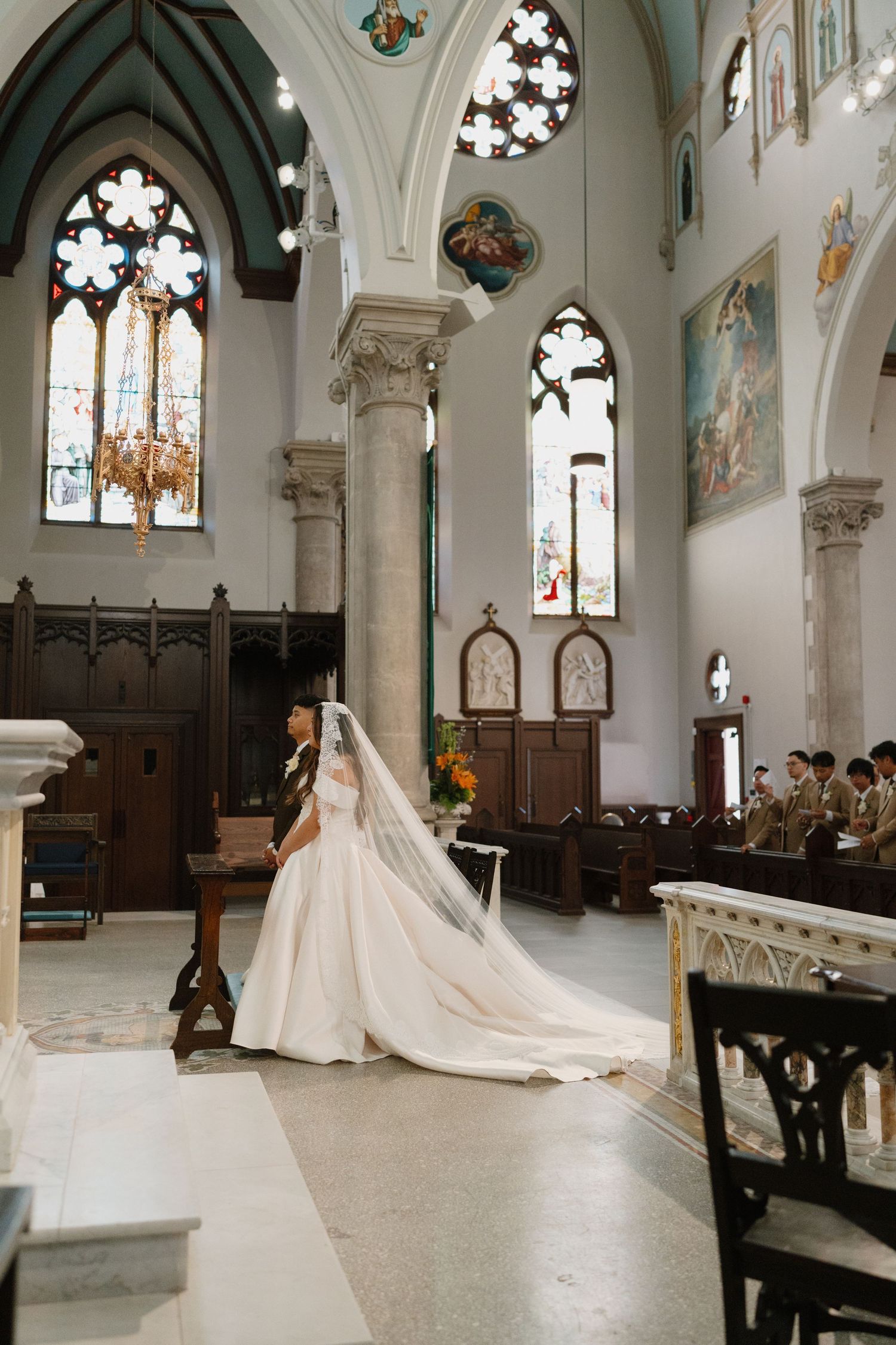 Bride in elegant white gown with long veil kneeling at church altar during wedding ceremony with stained glass windows.