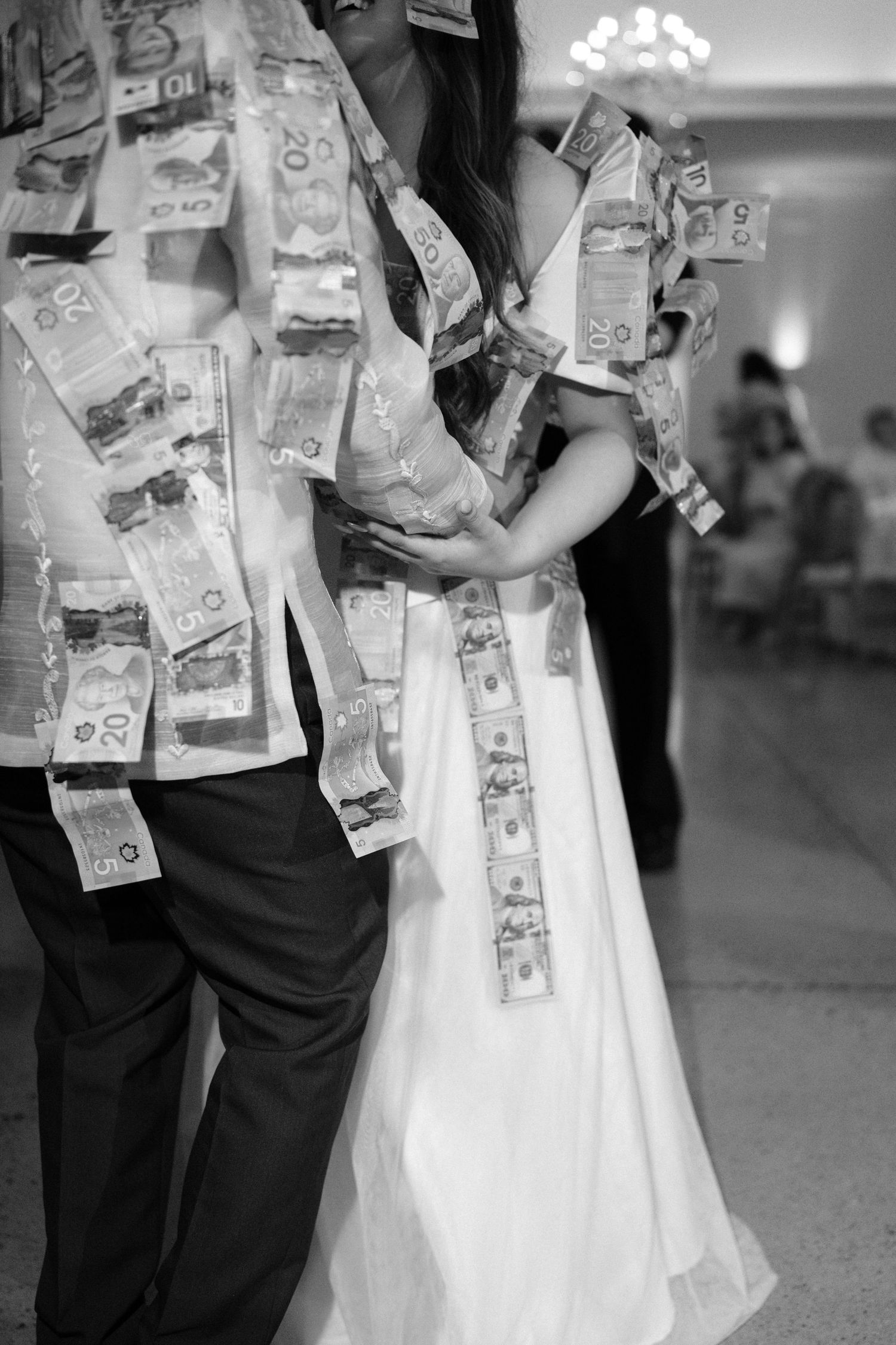 Black and white photo of a bride and groom dancing, covered in pinned paper currency bills at a wedding money dance.