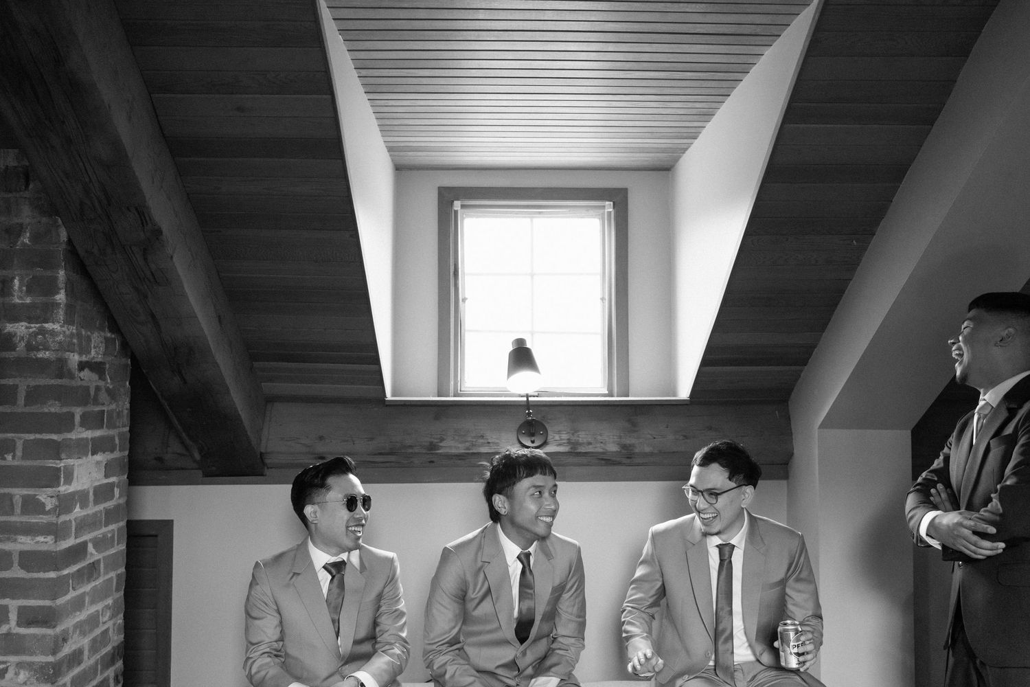 Three groomsmen in suits sit together on a bench in a rustic venue, laughing and relaxing before a wedding ceremony.