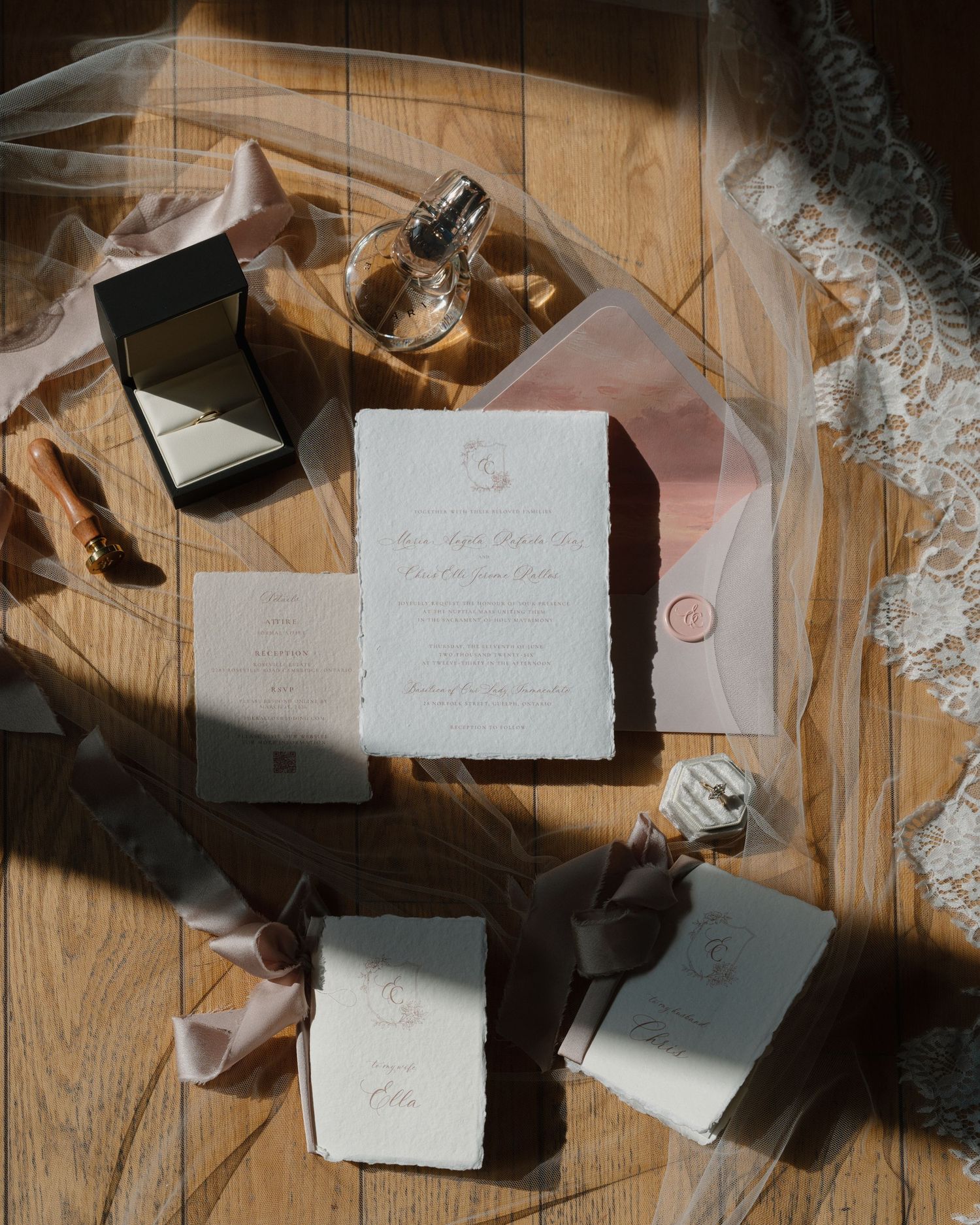 Elegant wedding invitation suite with deckled edges, pink envelope, perfume, ribbon, and lace veil on wooden floor.