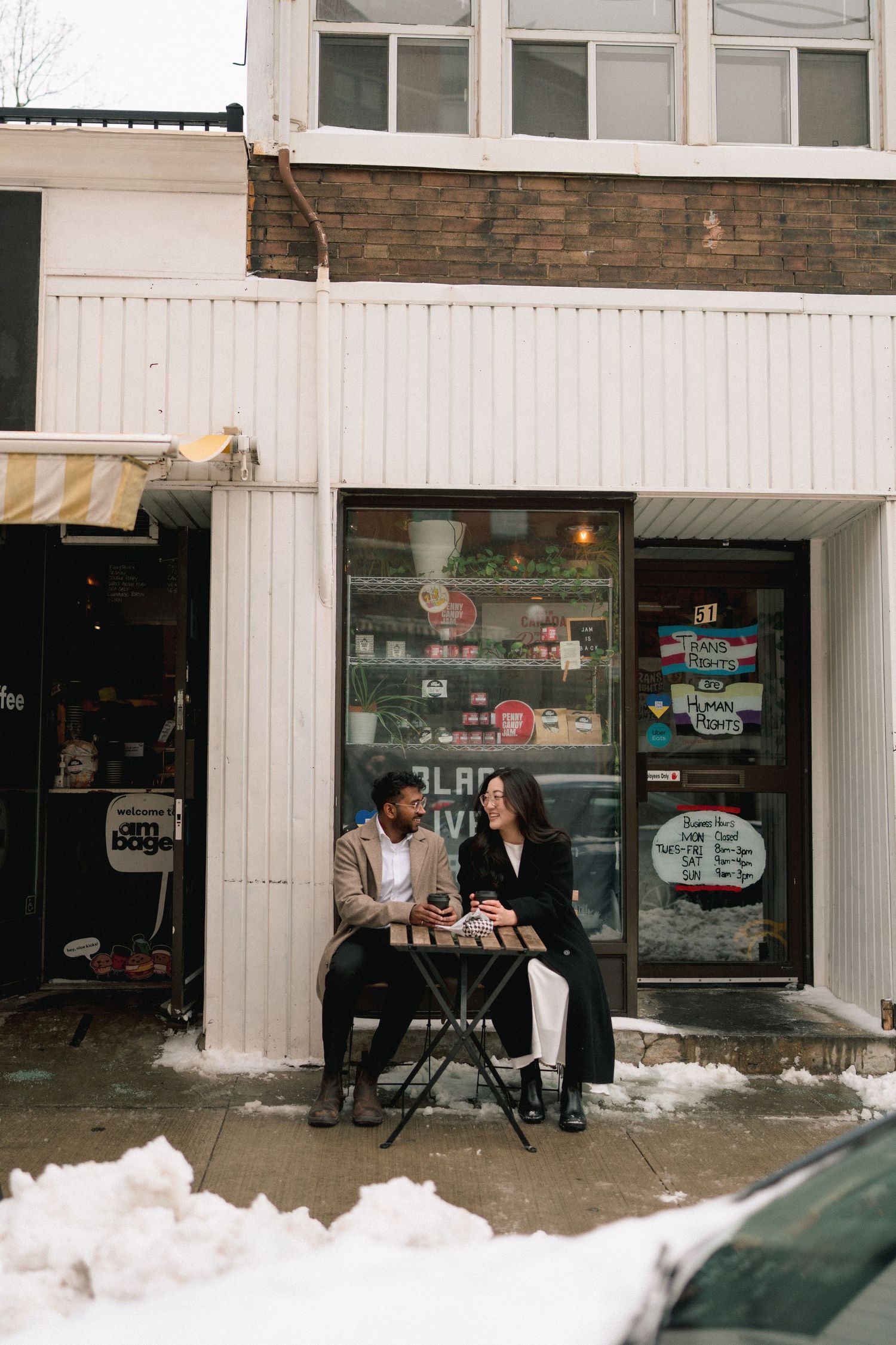 Two people sit at a table outside a storefront in winter with snow piled on the ground.