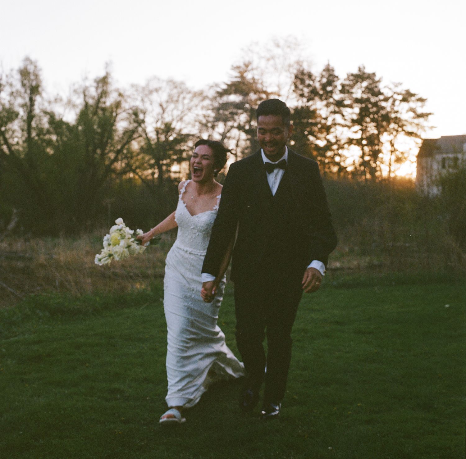 A joyful couple runs together on a grassy lawn at sunset during their wedding celebration.